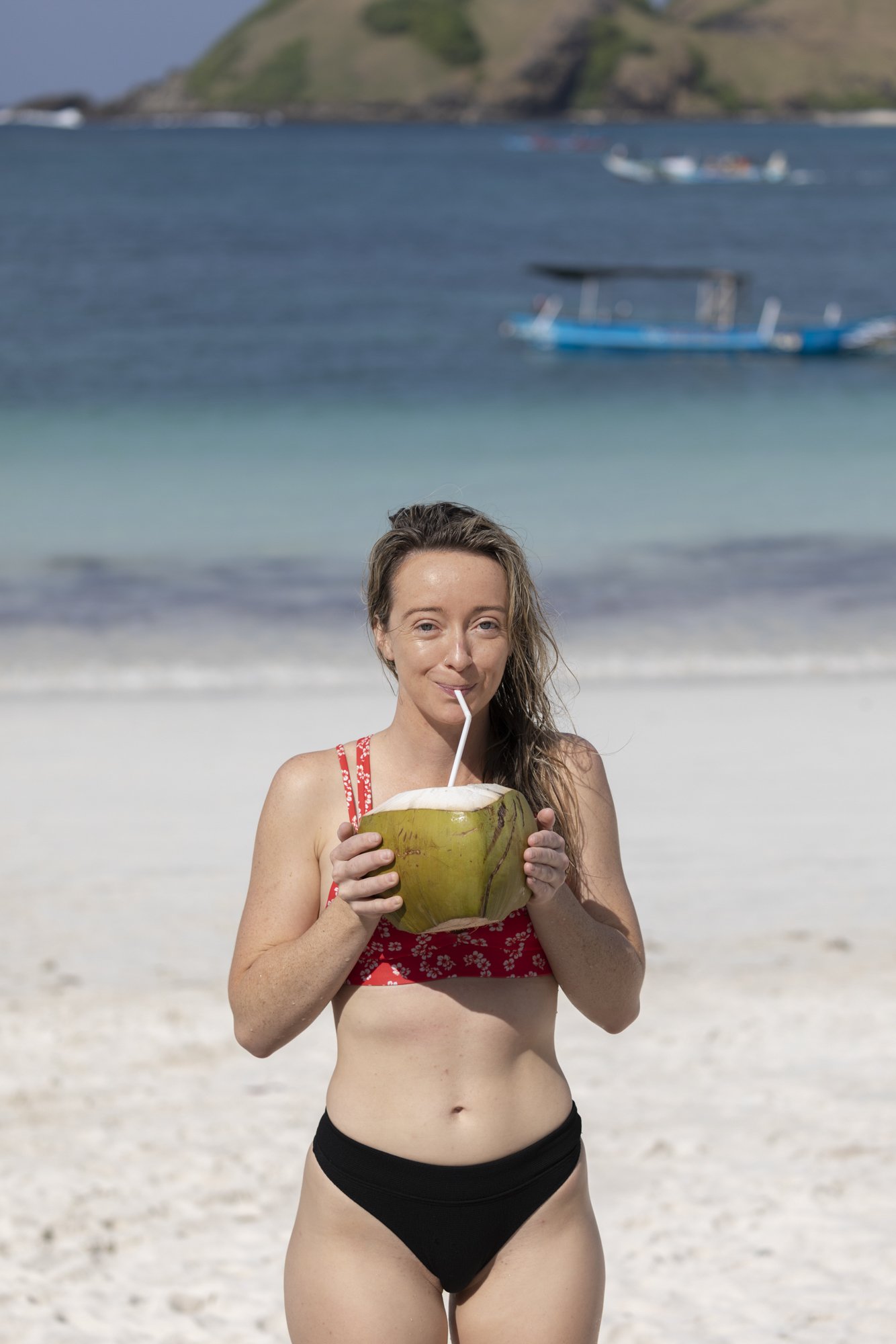 Woman in a red bikini top and black bottoms standing on a sandy beach, drinking from a coconut with a straw. The ocean with boats and green hills are in the background.