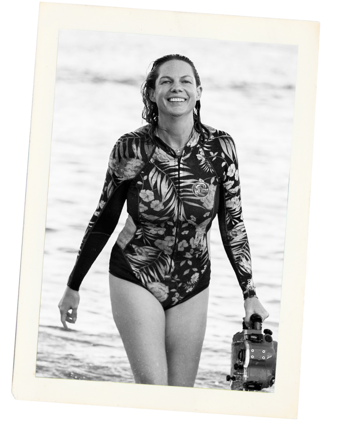 Black and white photo of a smiling woman in a floral wetsuit, holding a camera, walking in the water at the beach.