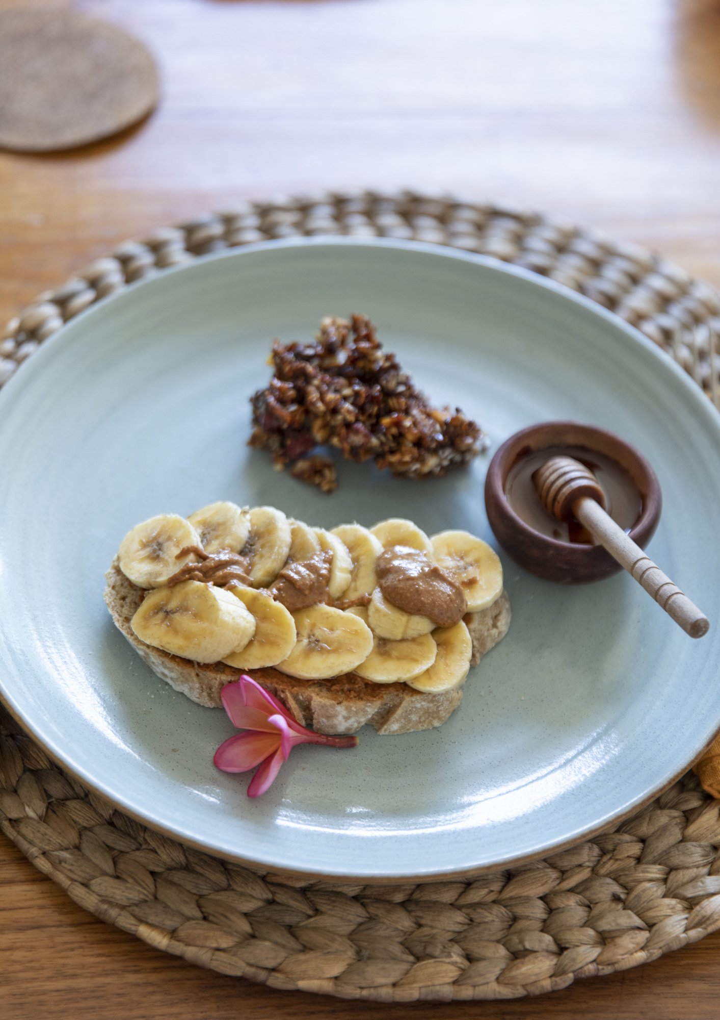 Slice of bread topped with sliced bananas and peanut butter, a serving of granola, a small bowl of honey with a wooden honey dipper, and a pink flower on a light blue plate set on a woven placemat.