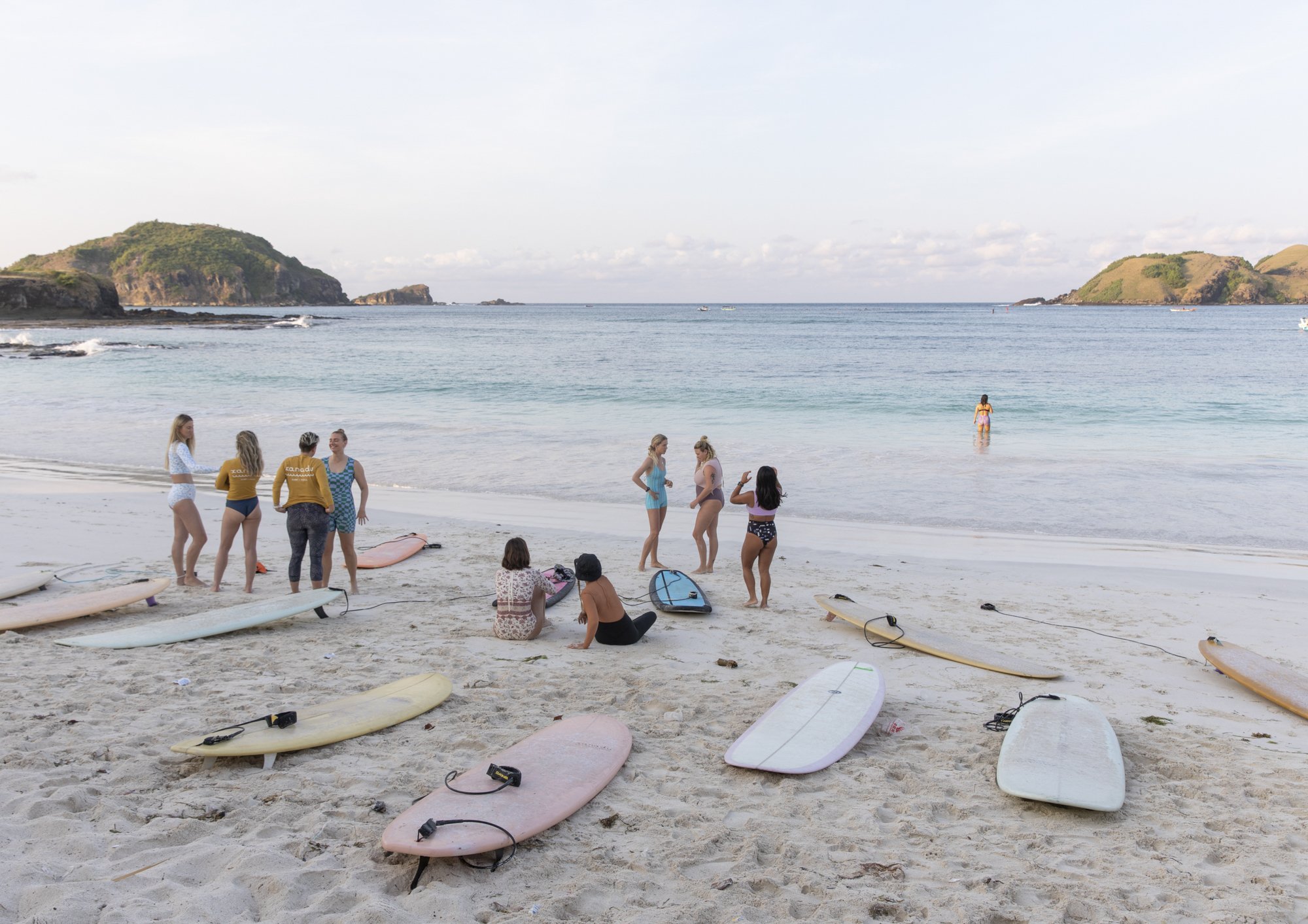 Beach scene with several surfboards laid on the sand, a group of women standing and sitting near the shoreline, some in swimsuits, engaging in conversation, and others in the water, with distant hills and calm ocean in the background.