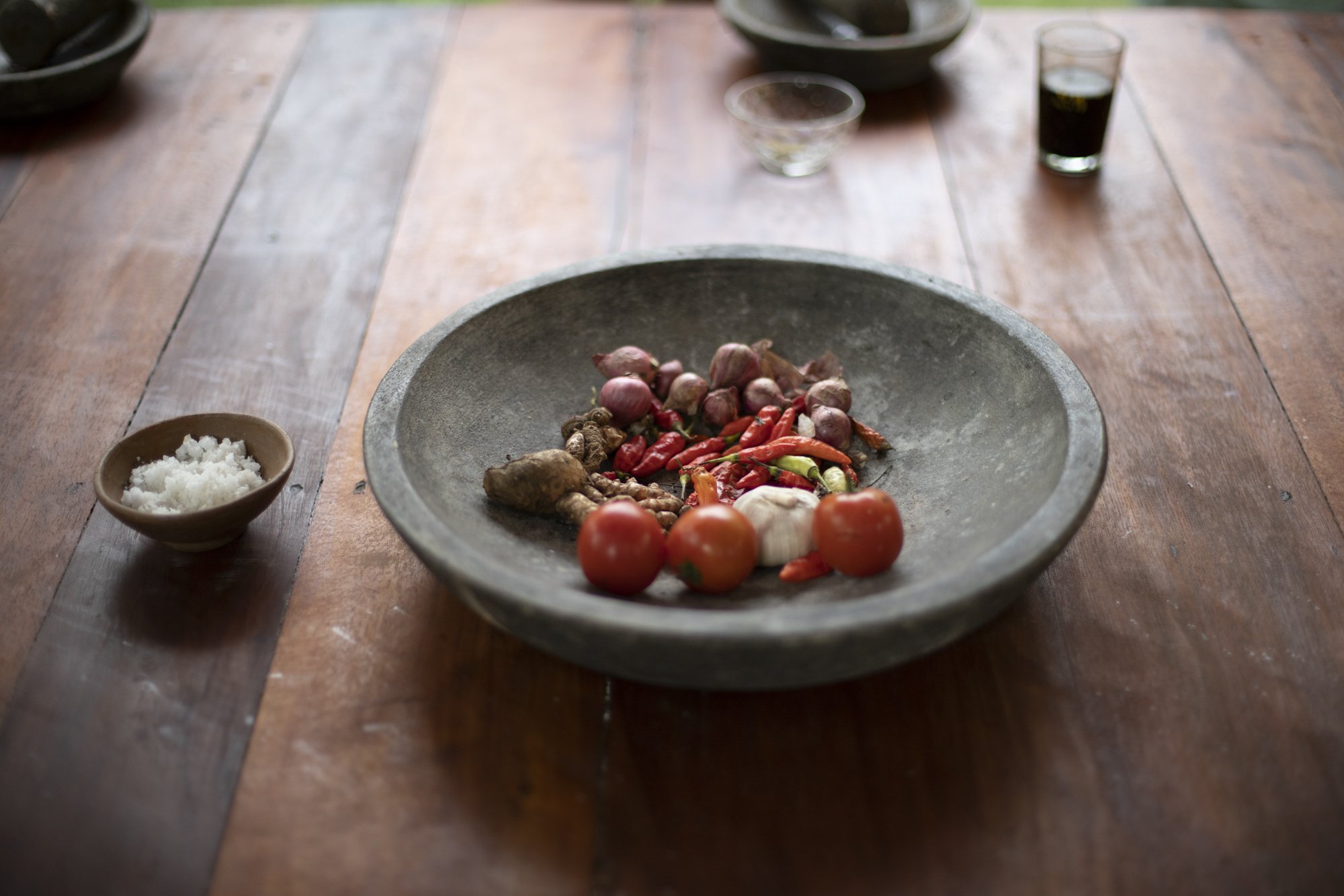 A large gray bowl with various fresh vegetables including tomatoes, garlic, red onions, and chili peppers, on a wooden table. Smaller bowls with salt and drinks are also visible in the background.