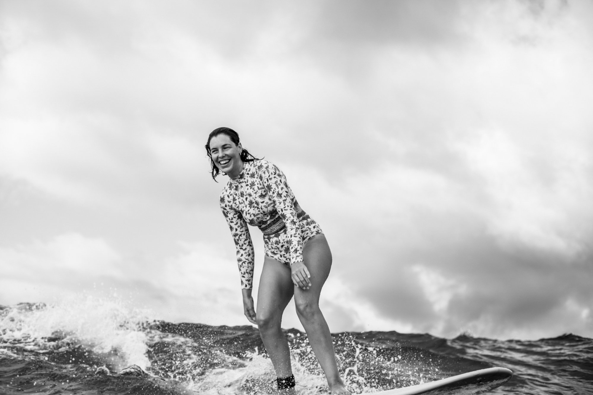 A woman in a patterned long-sleeve top and shorts surfing on a wave, smiling, in black and white.