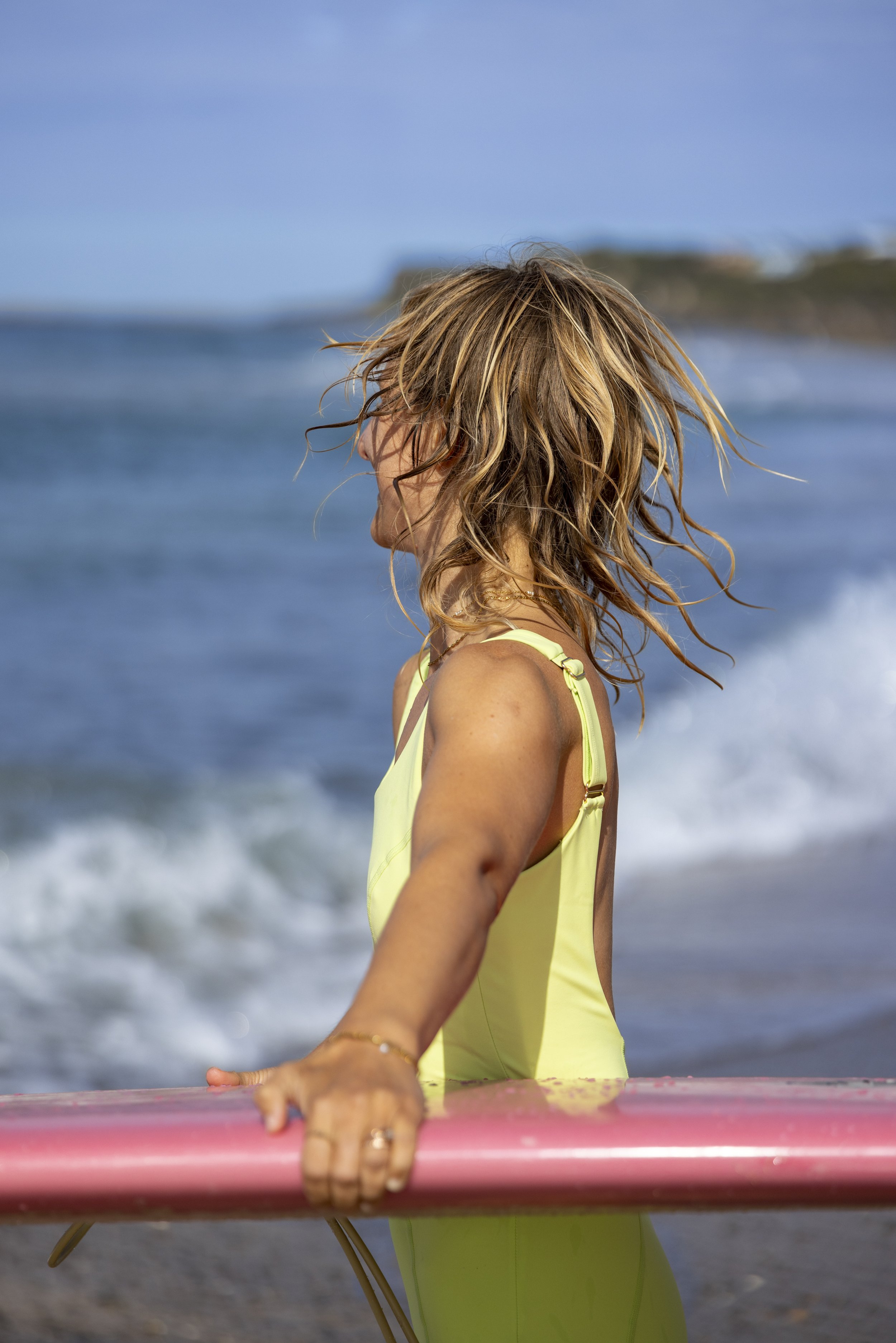 Woman with blonde hair wearing a yellow swimsuit holding a pink surfboard at the beach with ocean waves in the background.