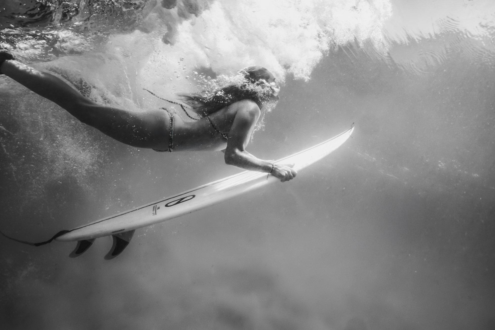 Black and white underwater photo of a person holding a surfboard, submerged, with waves breaking above.