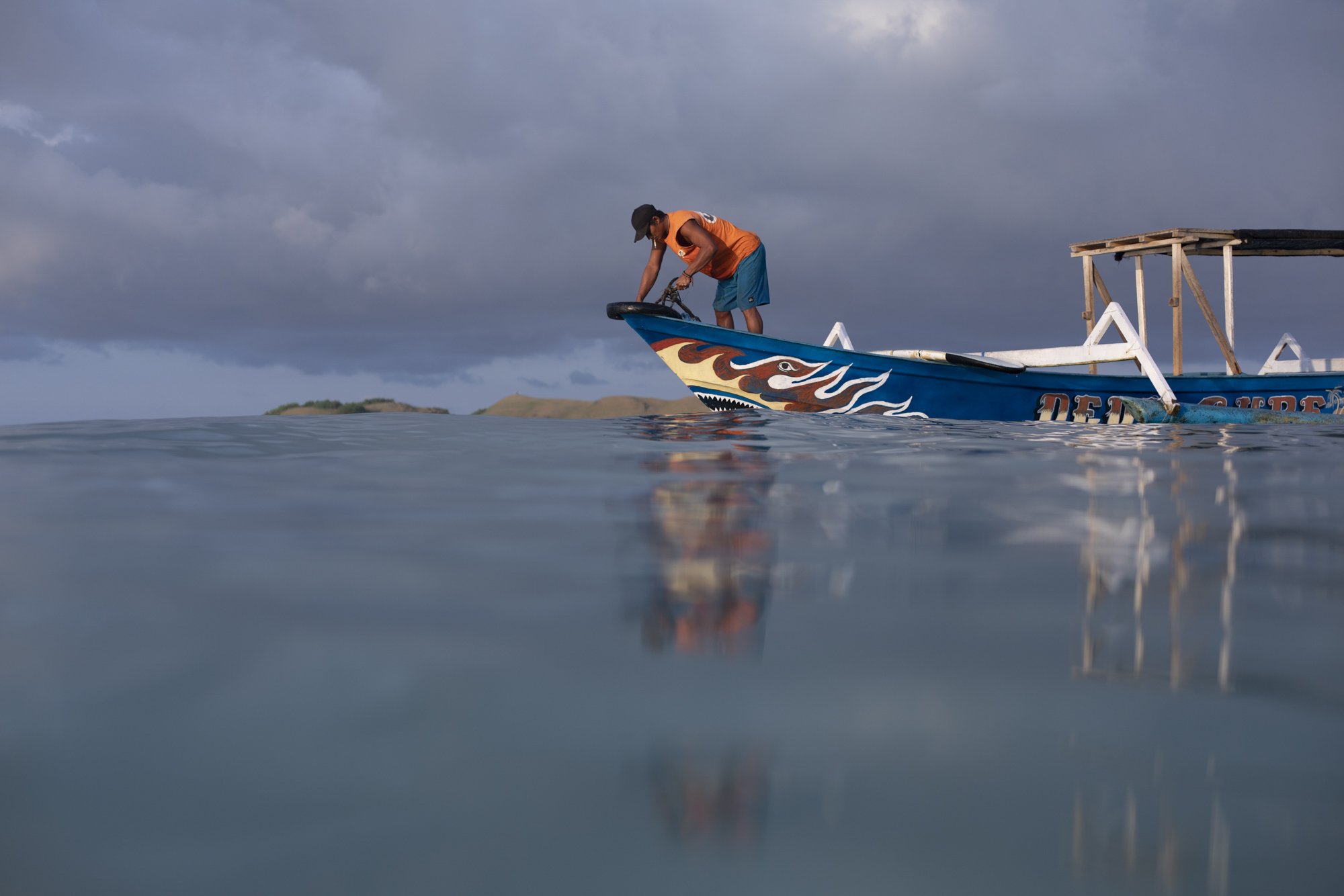 A person in an orange shirt and blue shorts stands on the edge of a decorative boat in calm water, preparing to paddle as dark storm clouds loom overhead.