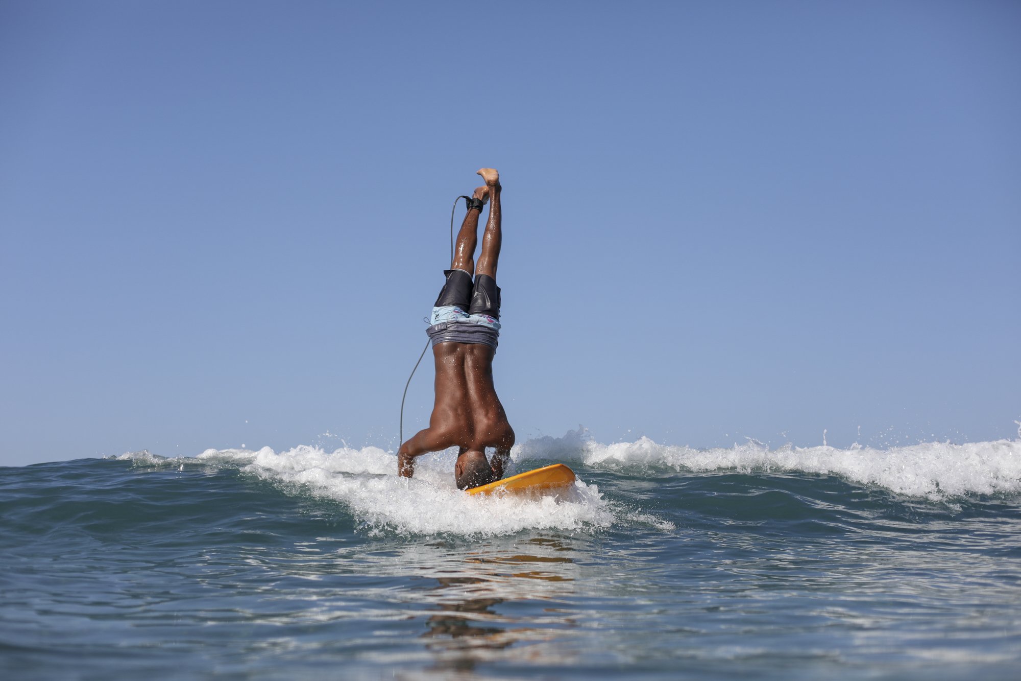 A person performing a handstand on a surfboard in the water at the beach with a clear blue sky.