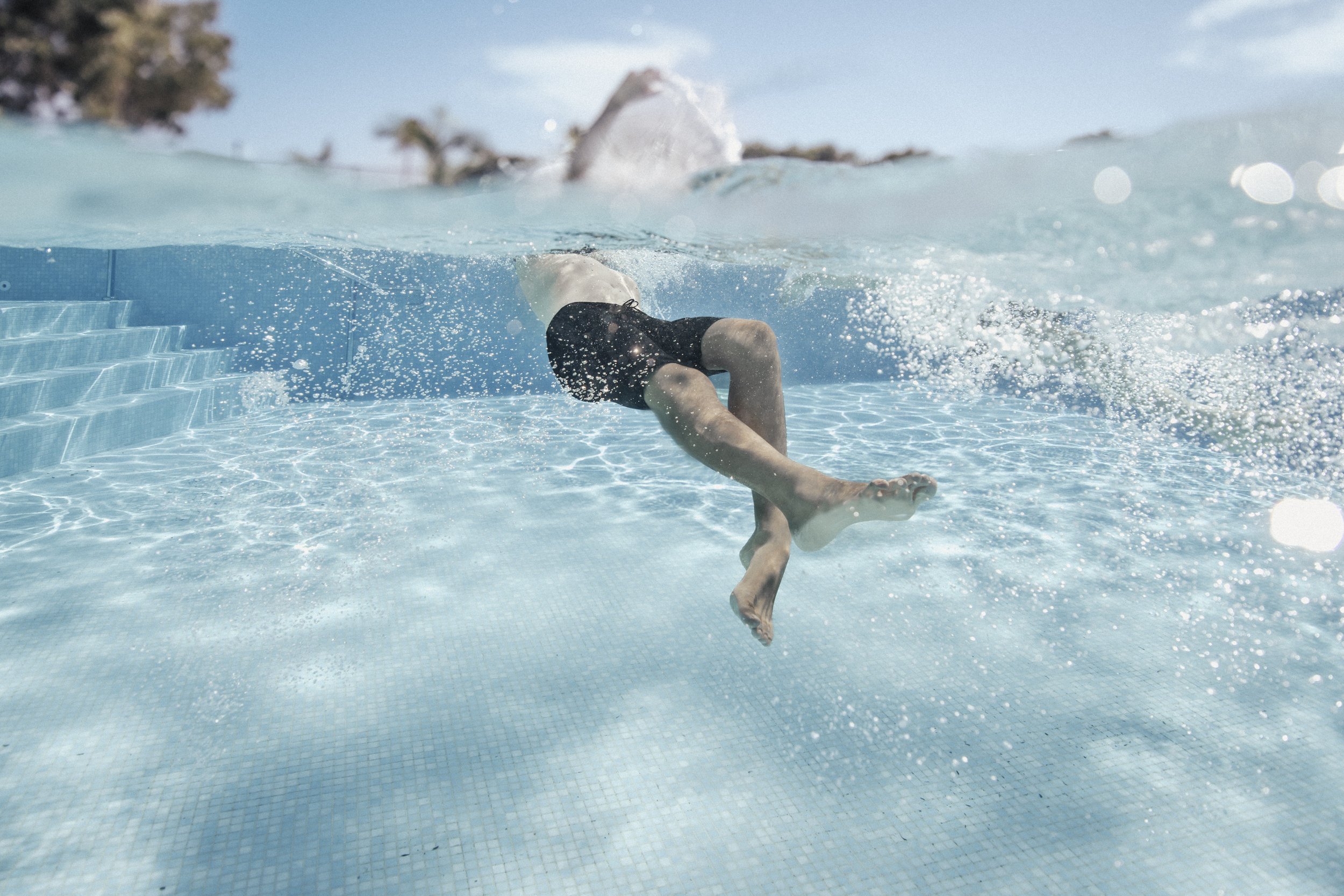 A child diving into a swimming pool from the water's surface, with a blue tile pool floor visible.