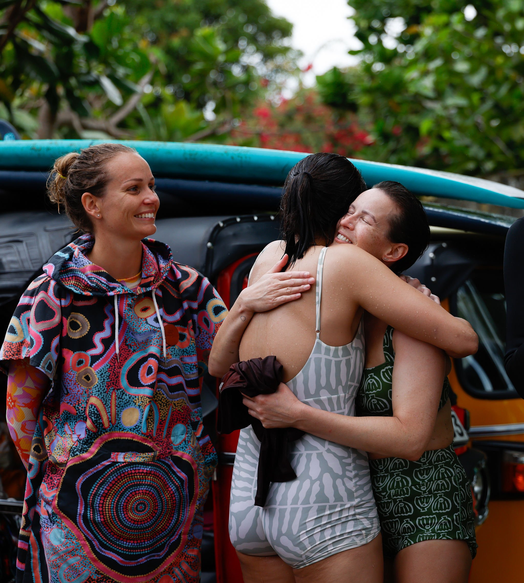 Three women embrace and smile after surfing, with surfboards on a vehicle and tropical trees in the background.