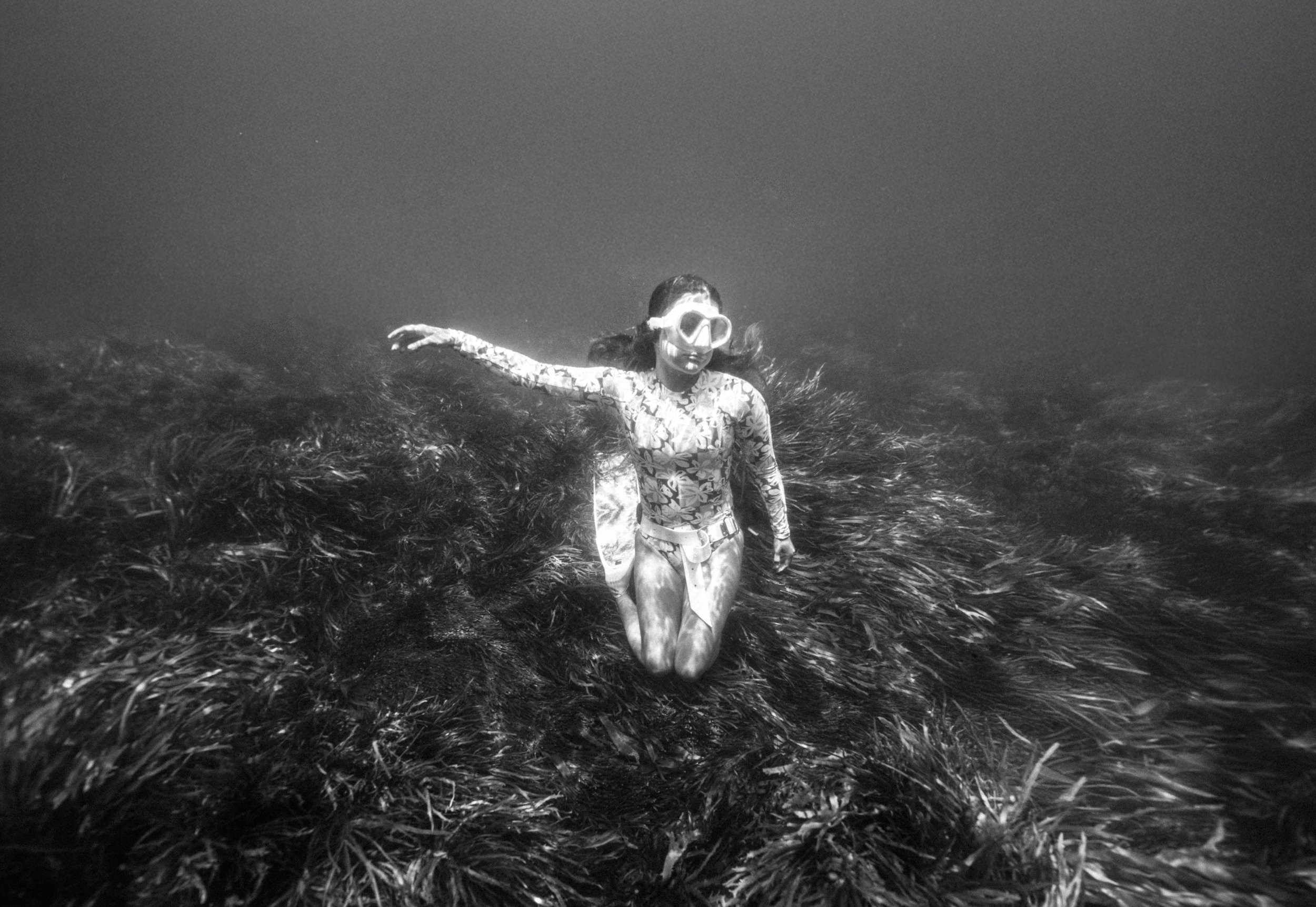A woman in snorkeling gear kneeling on a submerged rocky seafloor covered with seaweed, captured in black-and-white underwater photograph.
