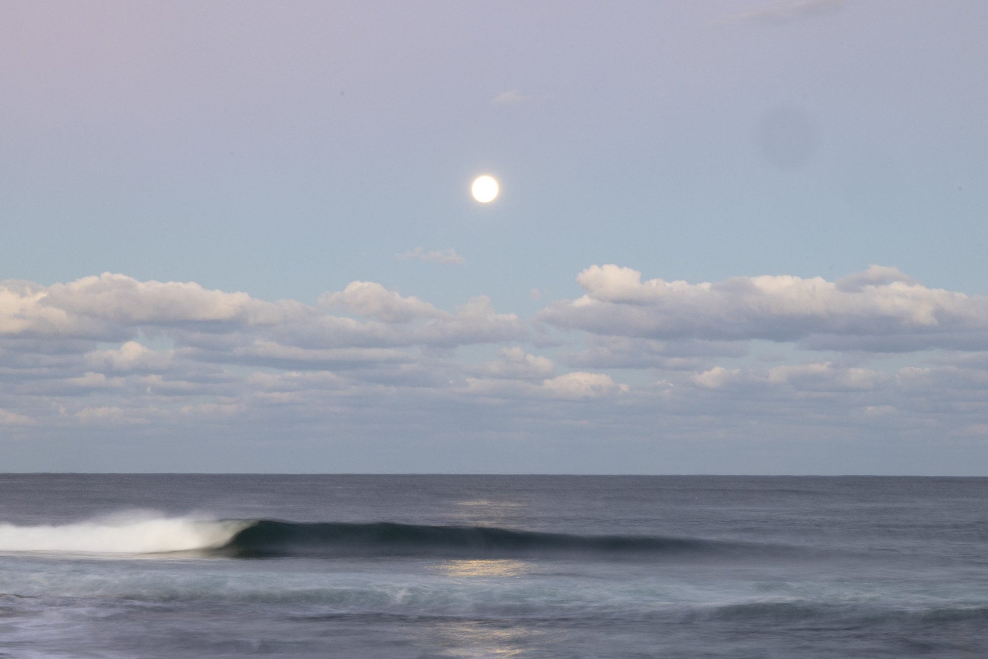 A calm ocean scene at dusk with a small wave, cloud-filled sky, and a bright full moon