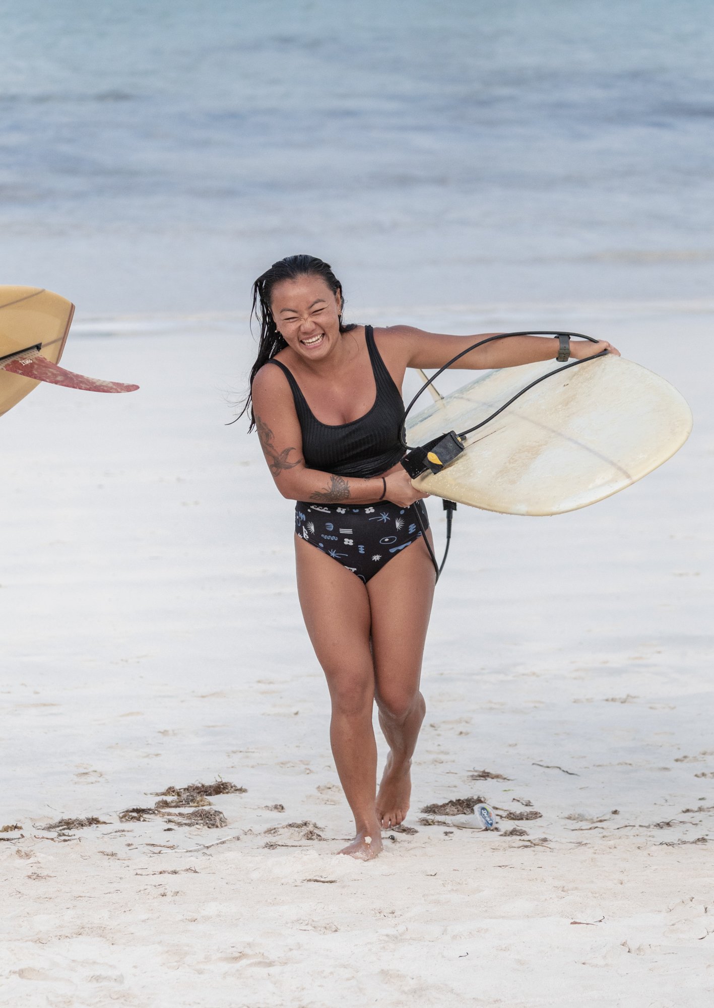 A woman in a black swimsuit with tattoos is smiling and holding a surfboard on the beach near the ocean.