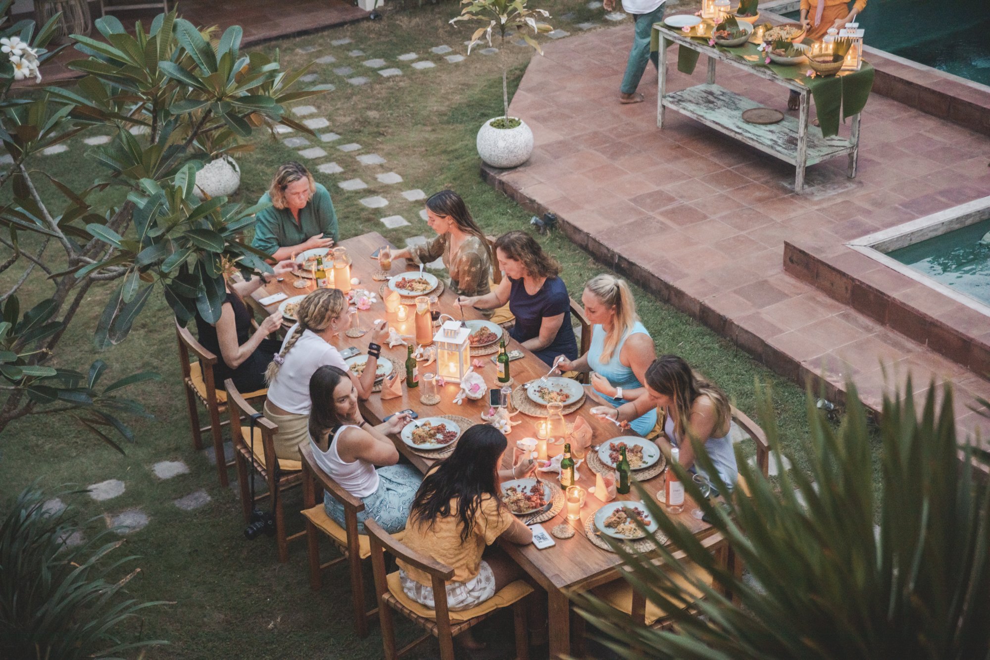 A group of women enjoying a dinner party at a long outdoor table set with plates of food, bottles of drinks, and candles, in a garden setting next to a pool.
