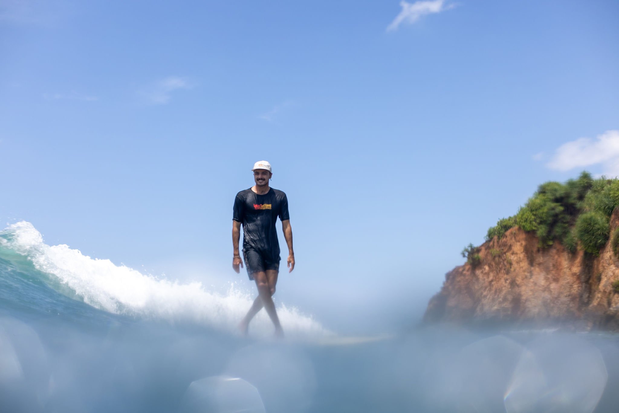 A man in a black t-shirt and white cap stands on a surfboard in the ocean, with a blue sky and green hillside in the background.