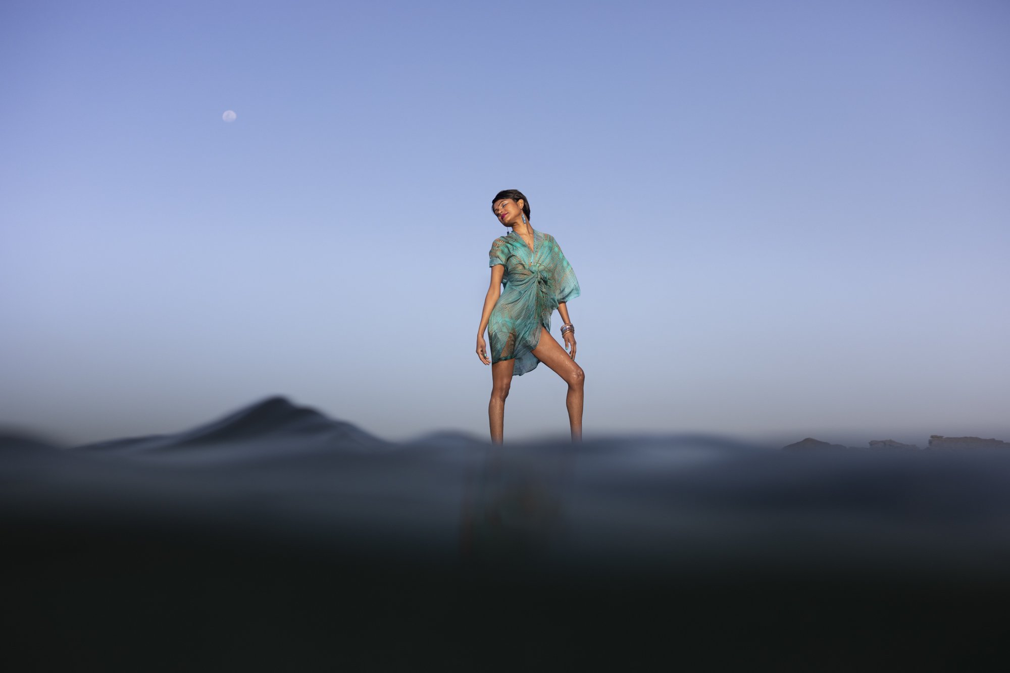 Woman in a sheer turquoise dress standing in shallow water with a clear sky and a visible moon in the background.