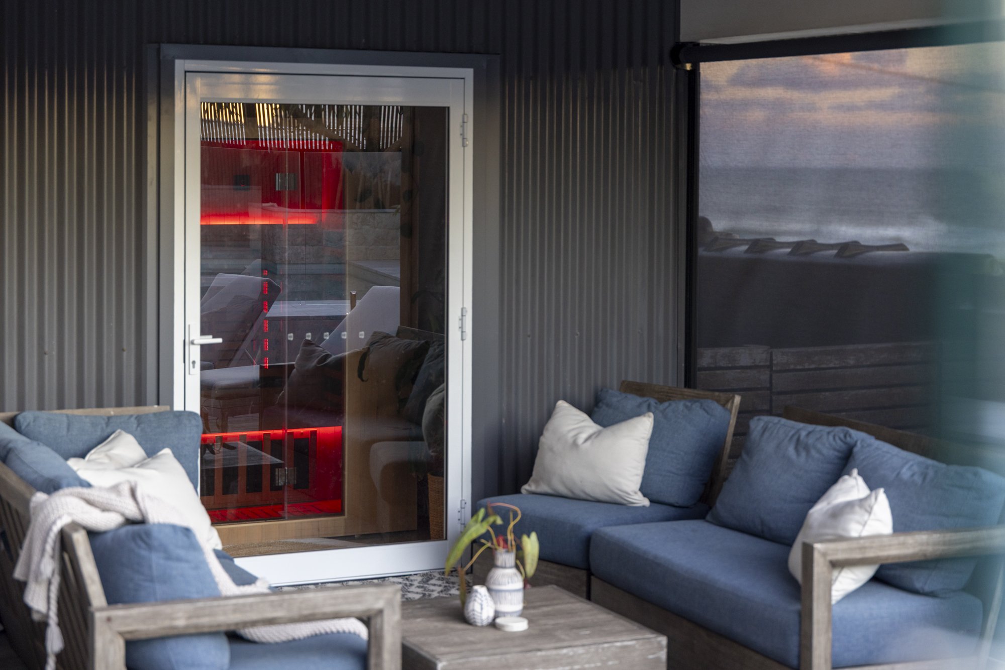 Outdoor patio with blue cushioned seating and pillows, a small wooden table with vases, and a black corrugated metal wall with a glass door reflecting red interior lights and an ocean view in the background.