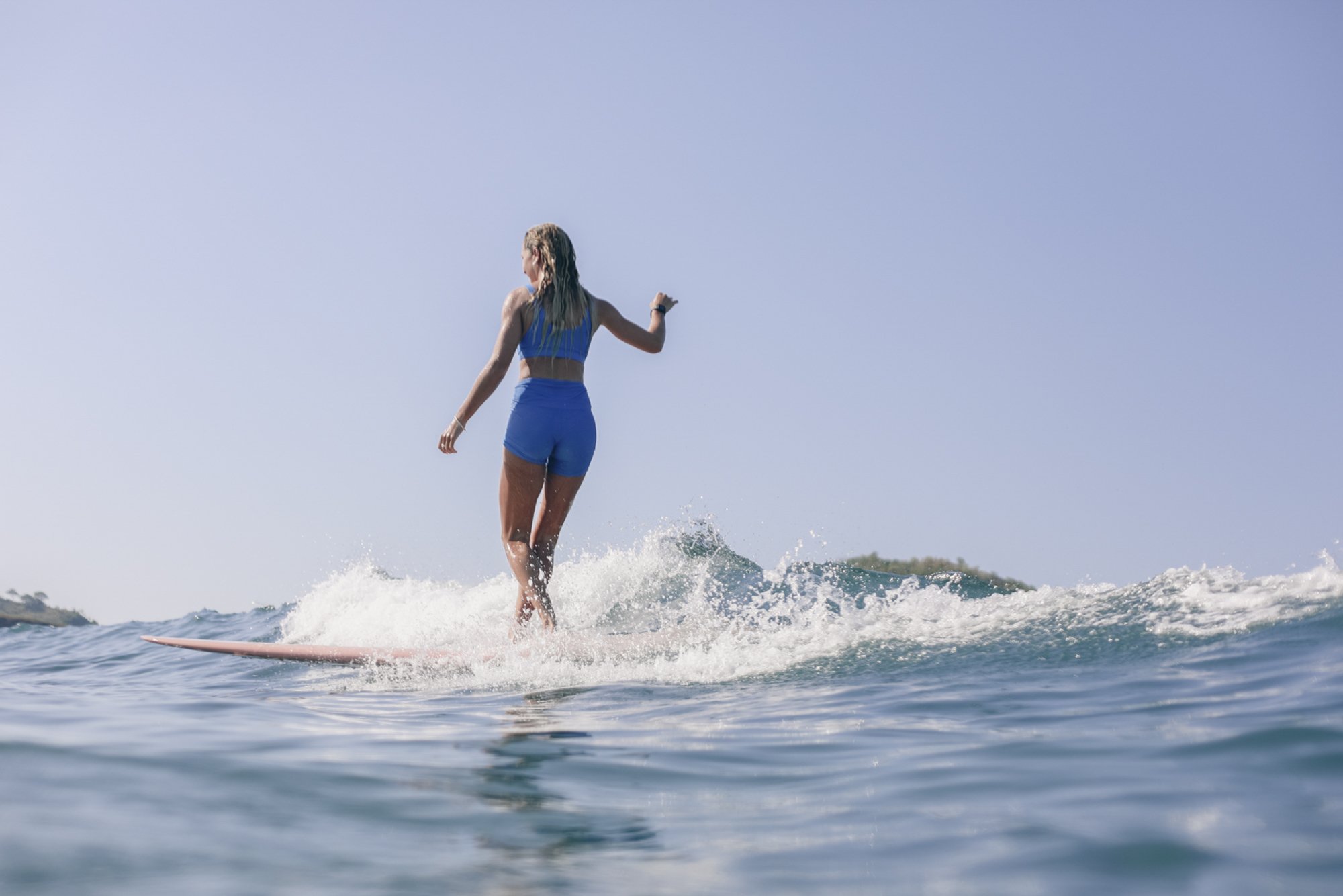 A woman surfing on a pink surfboard in the ocean, wearing a blue sports bra and matching shorts, with a clear blue sky above.
