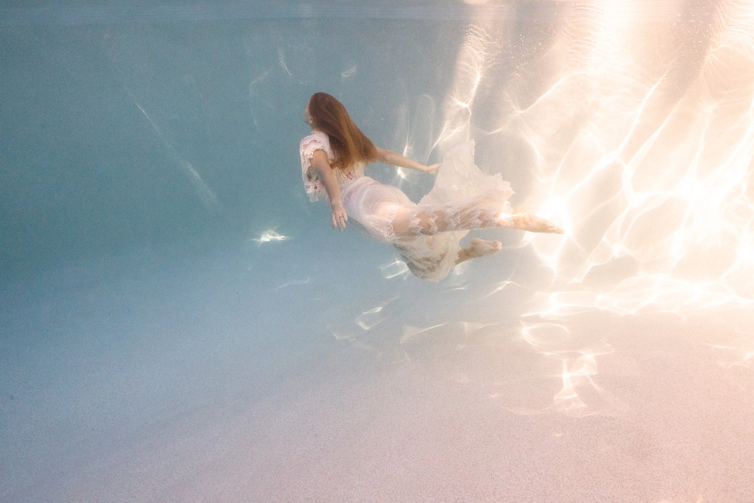 A woman with long red hair wearing a white dress is submerged and floating in water, with sunlight creating reflections on the water's surface.