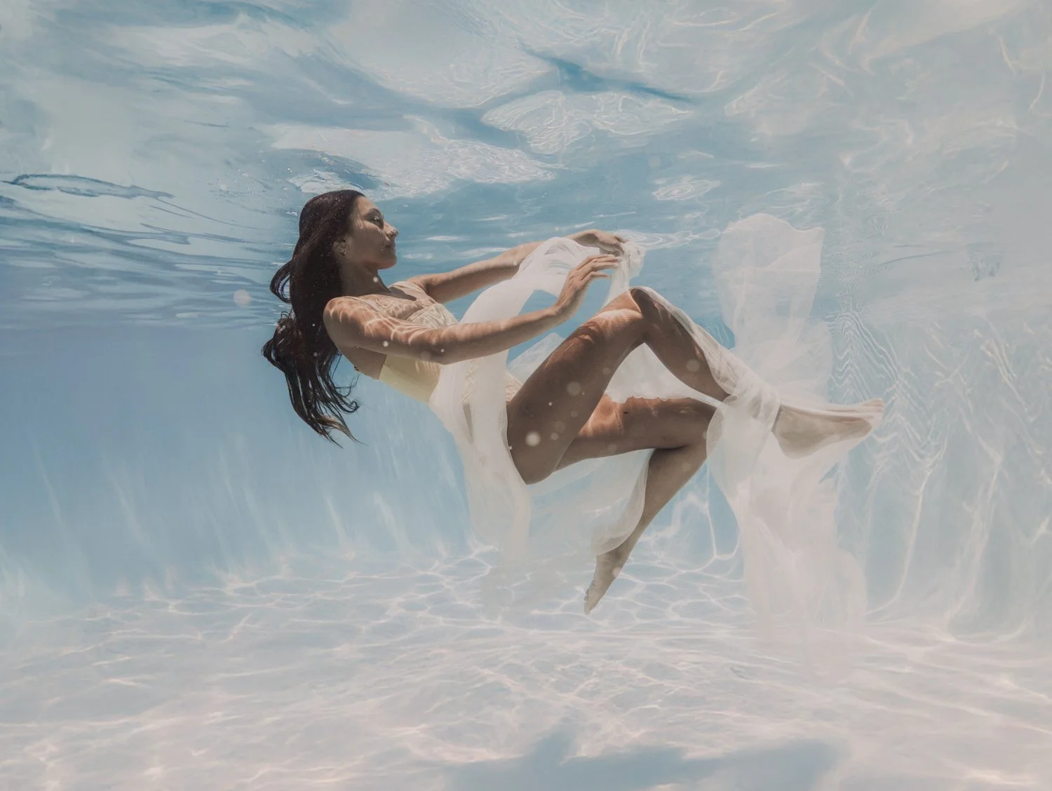 A woman in a light-colored dress floating underwater with her eyes closed and arms outstretched.
