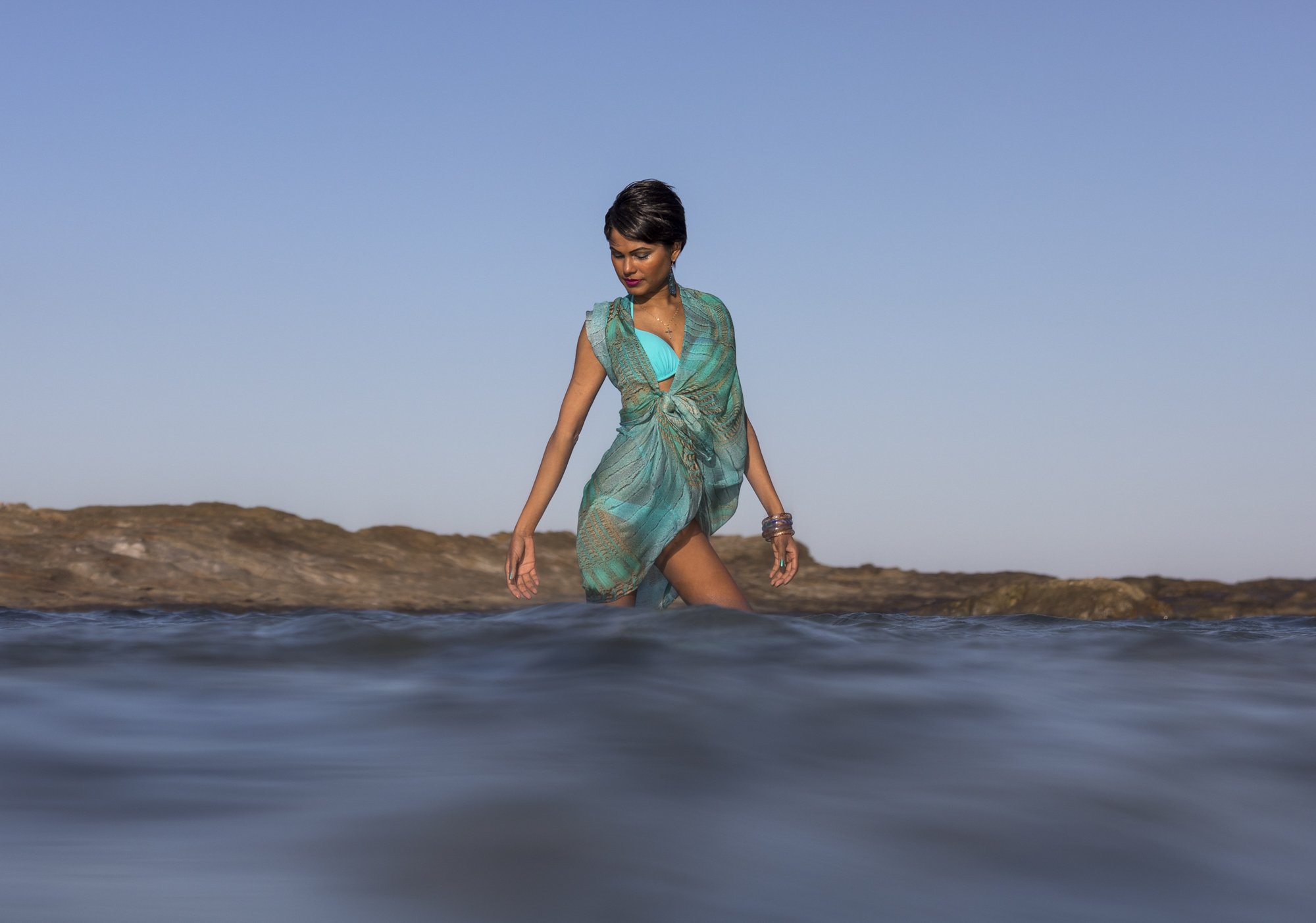 Woman in a turquoise swimsuit and sheer cover-up wading through shallow ocean water with rocky shoreline and clear sky in the background.