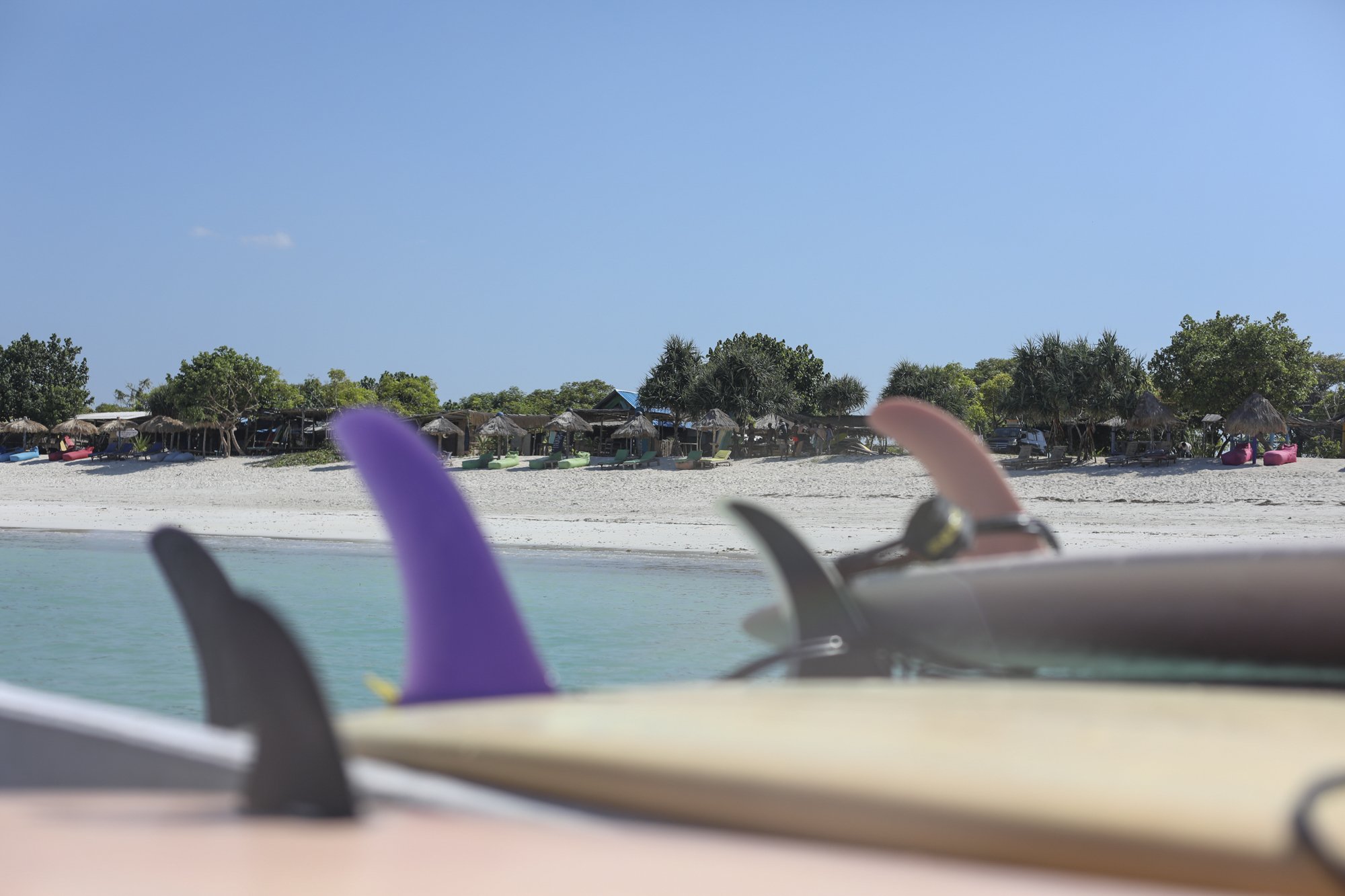 Colorful surfboards with fins resting on the beach, with a shoreline, trees, and beach huts in the background under a clear blue sky.