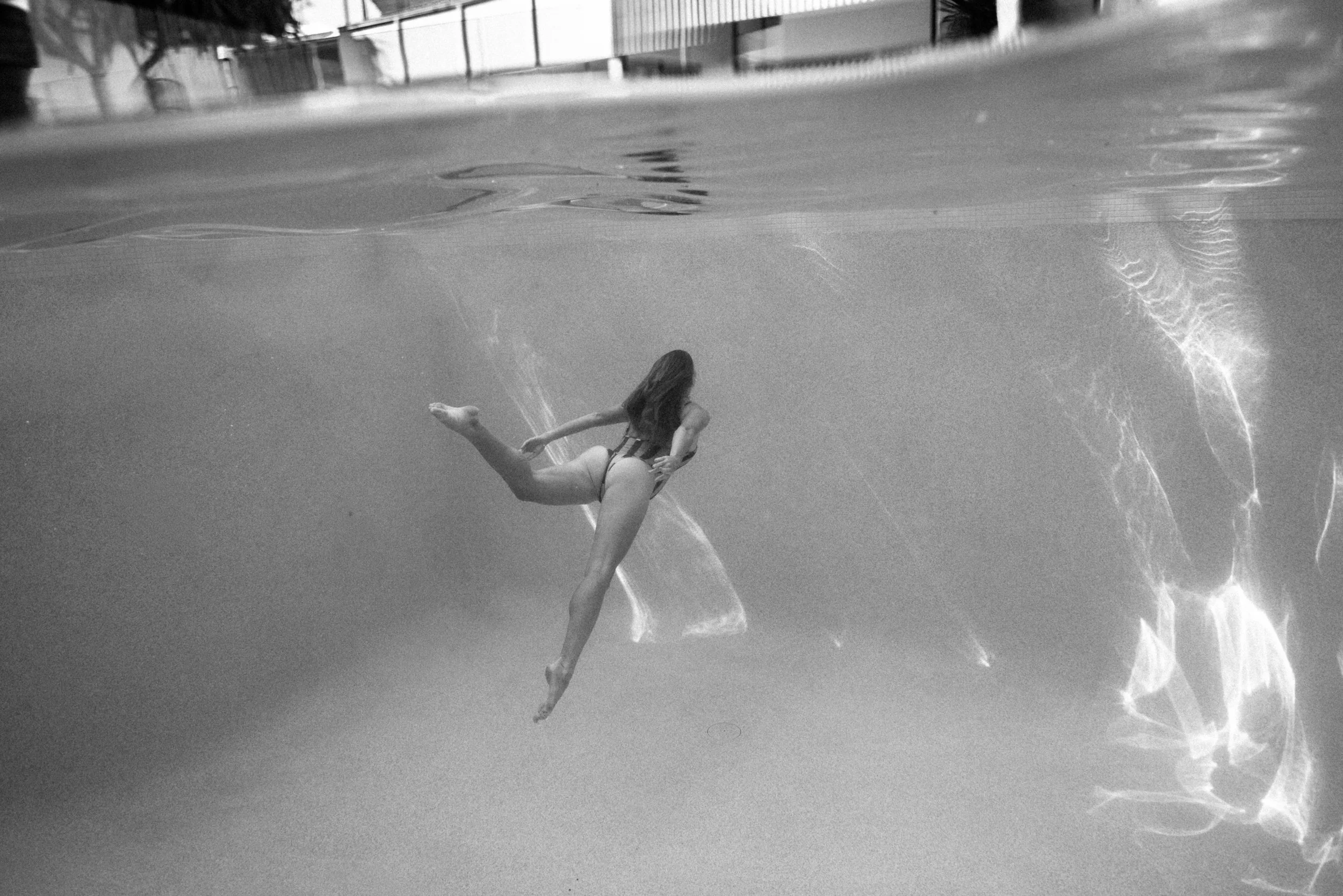 Black and white photo of a woman in a swimsuit swimming underwater in a pool, with reflections and light patterns on the water surface.