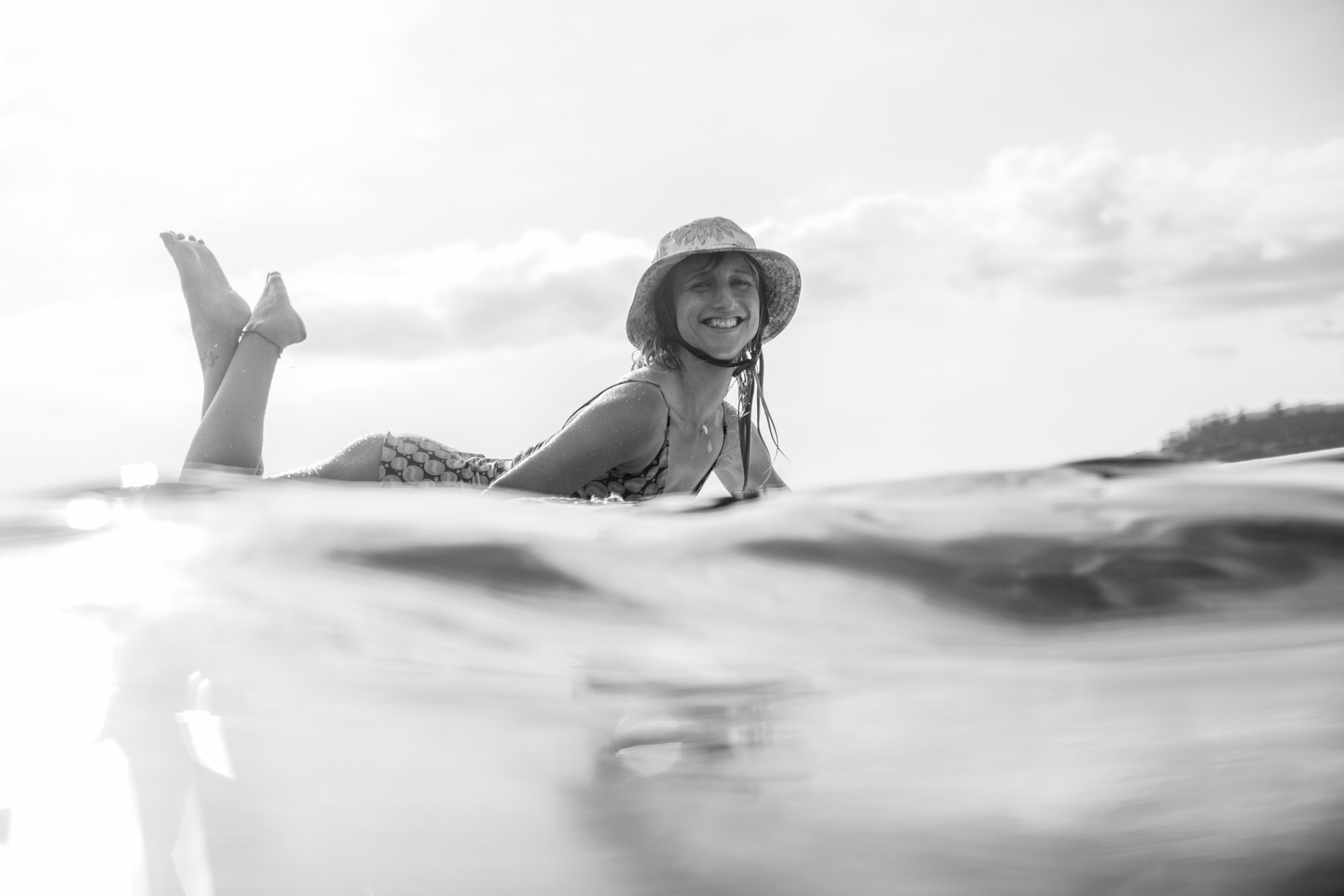 A woman smiling and lying on a surfboard in the ocean, wearing a hat and swimsuit, with a background of cloudy sky and distant land.