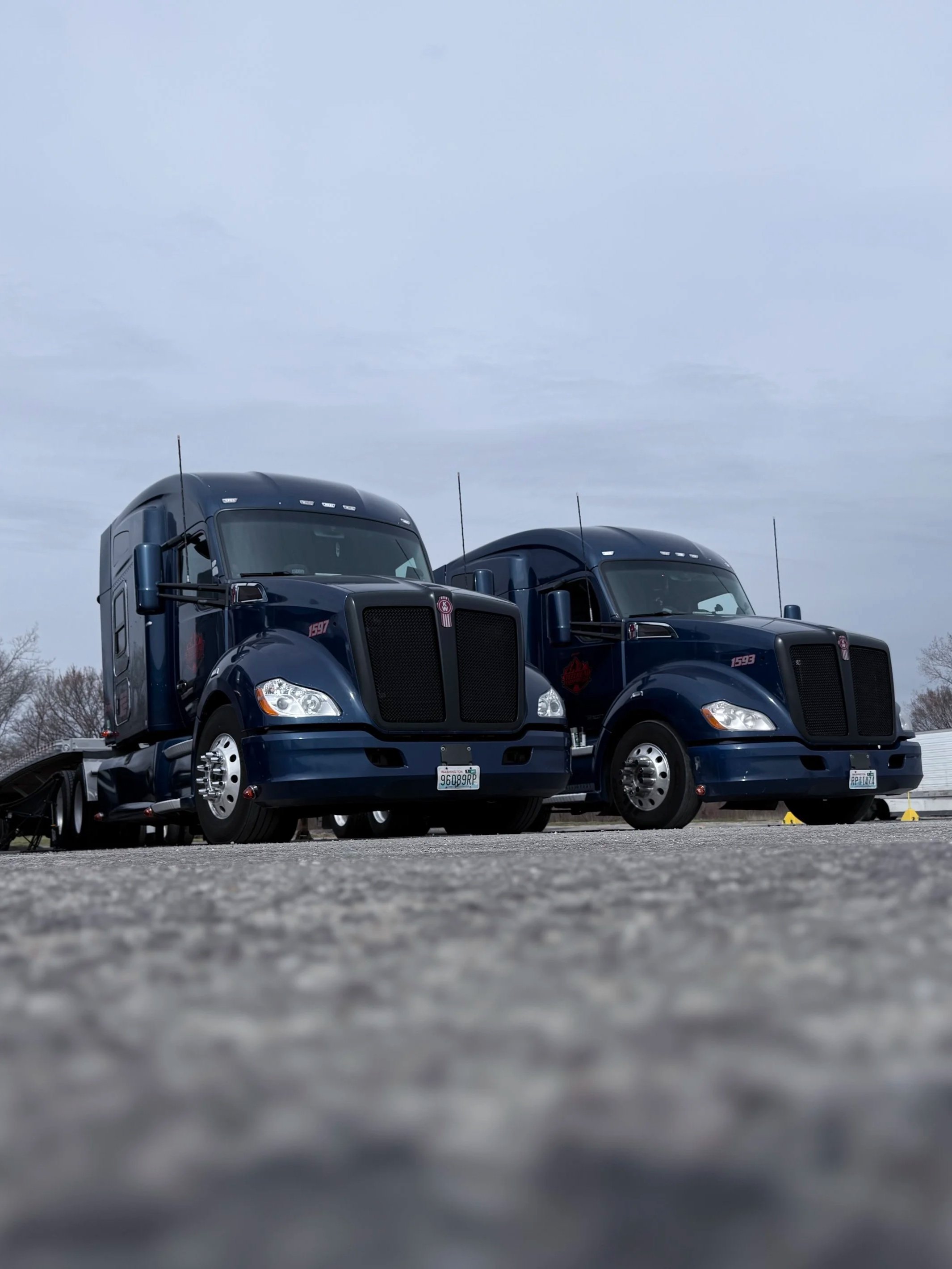 Two dark blue semi-trucks parked on a gravel lot under a cloudy sky during daytime, with some yellow cones visible in the background.