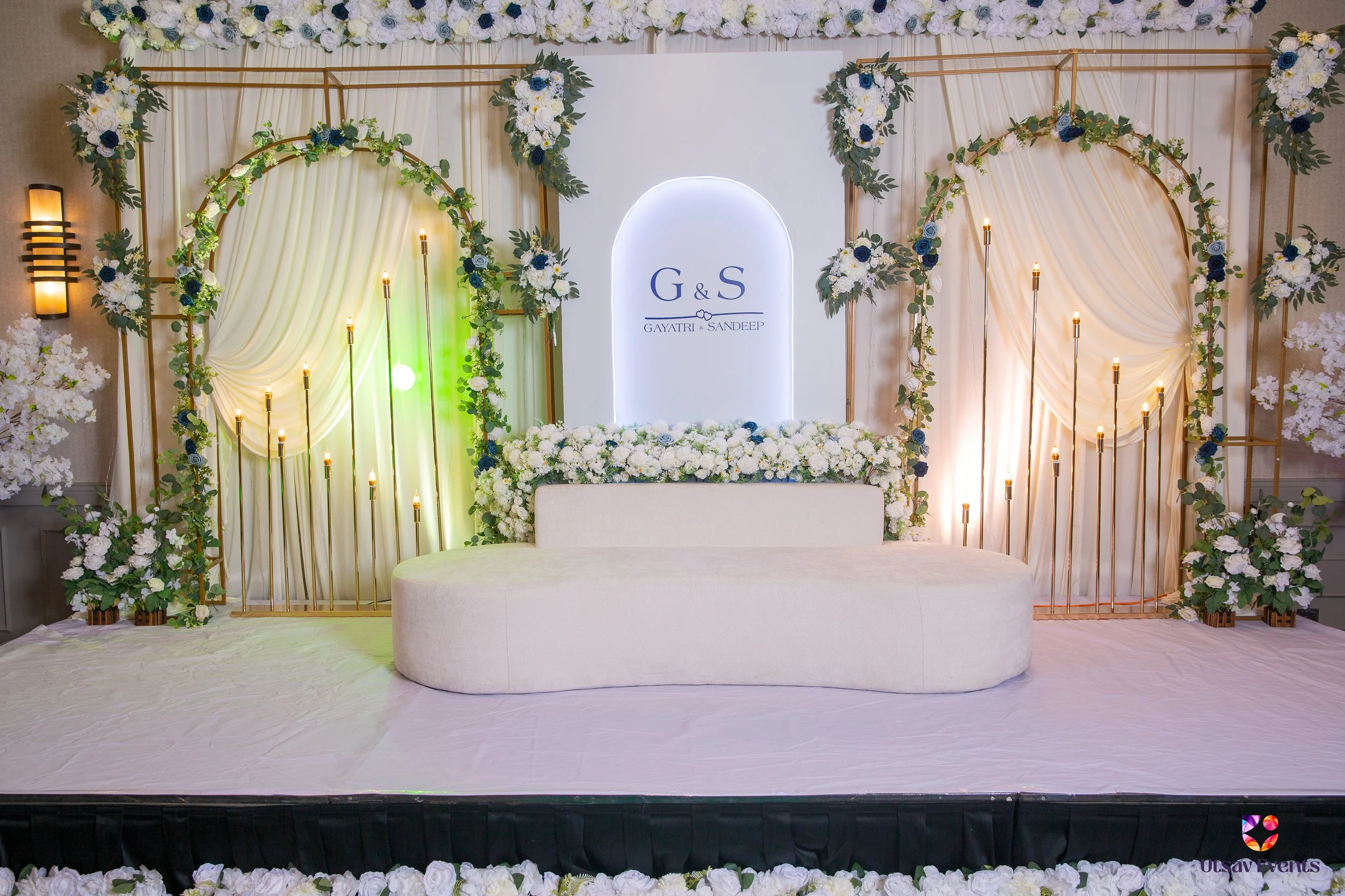 Wedding stage decorated with white flowers, blue accents, and gold accents, with draped white fabric, surrounded by floral arrangements, candles, and gold frames. A sign in the center reads 'G & S' with names 'Gayatri & Sandeep'.