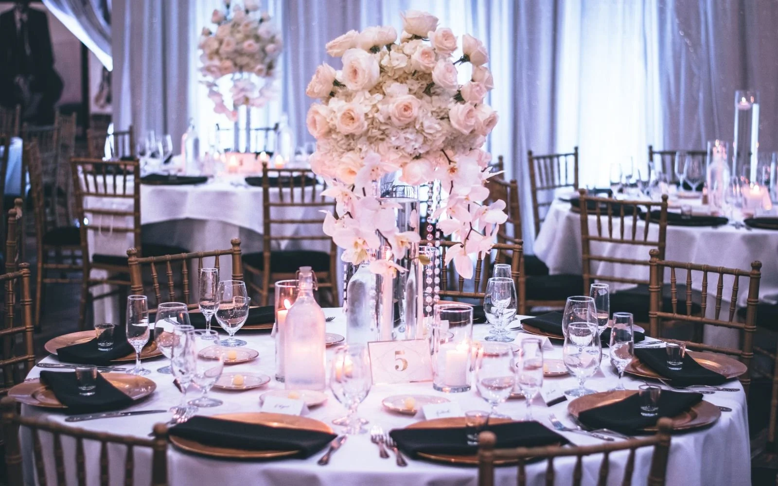 Elegant table setup with white linens, gold chargers, black napkins, and clear glassware, featuring a tall floral centerpiece of pink and white roses and orchids, in a decorated event space with draped curtains and dim lighting.