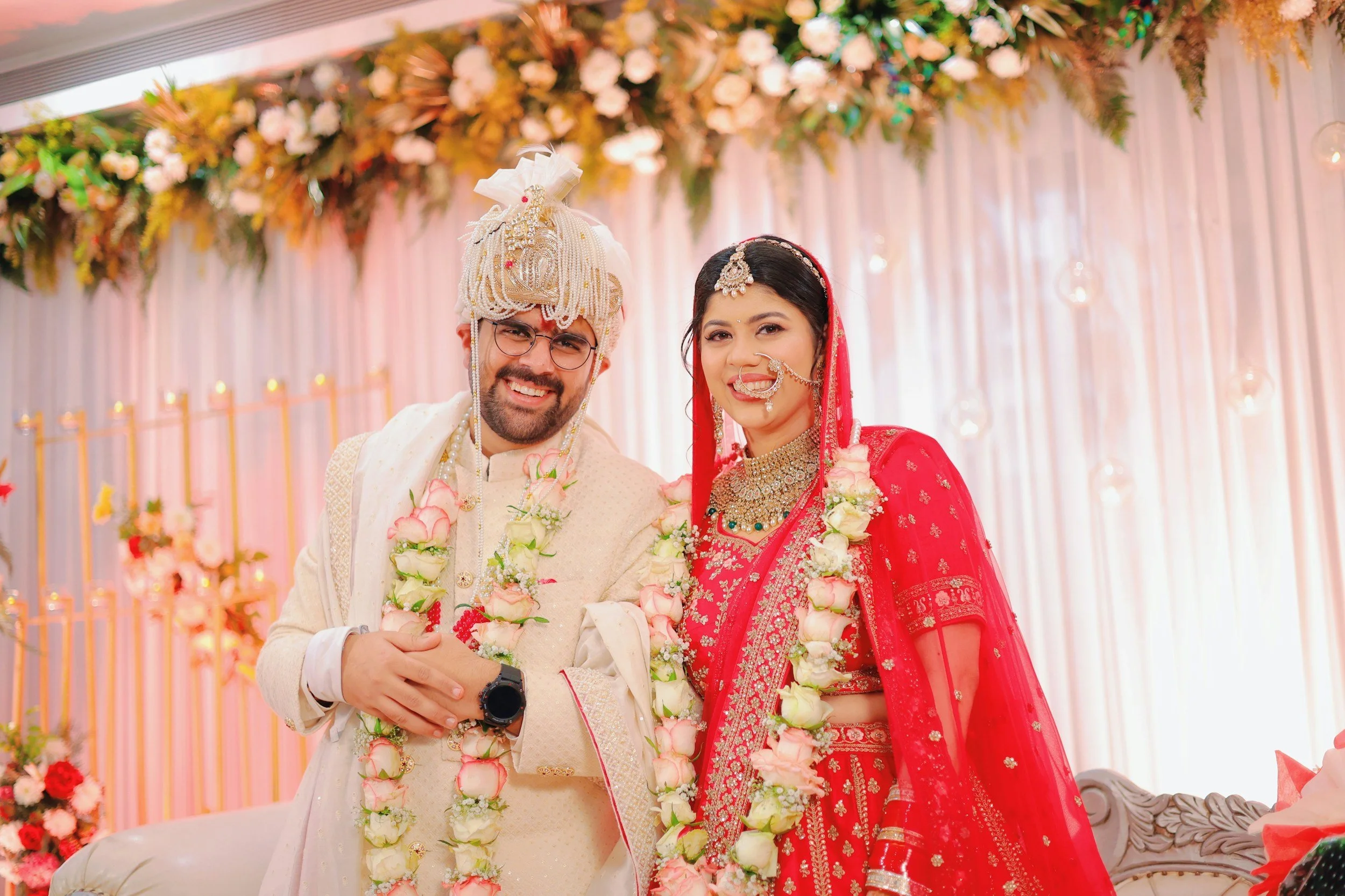 A smiling bride and groom in traditional Indian wedding attire are posing together at their wedding reception, with floral garlands around their necks and a decorated floral backdrop behind them.