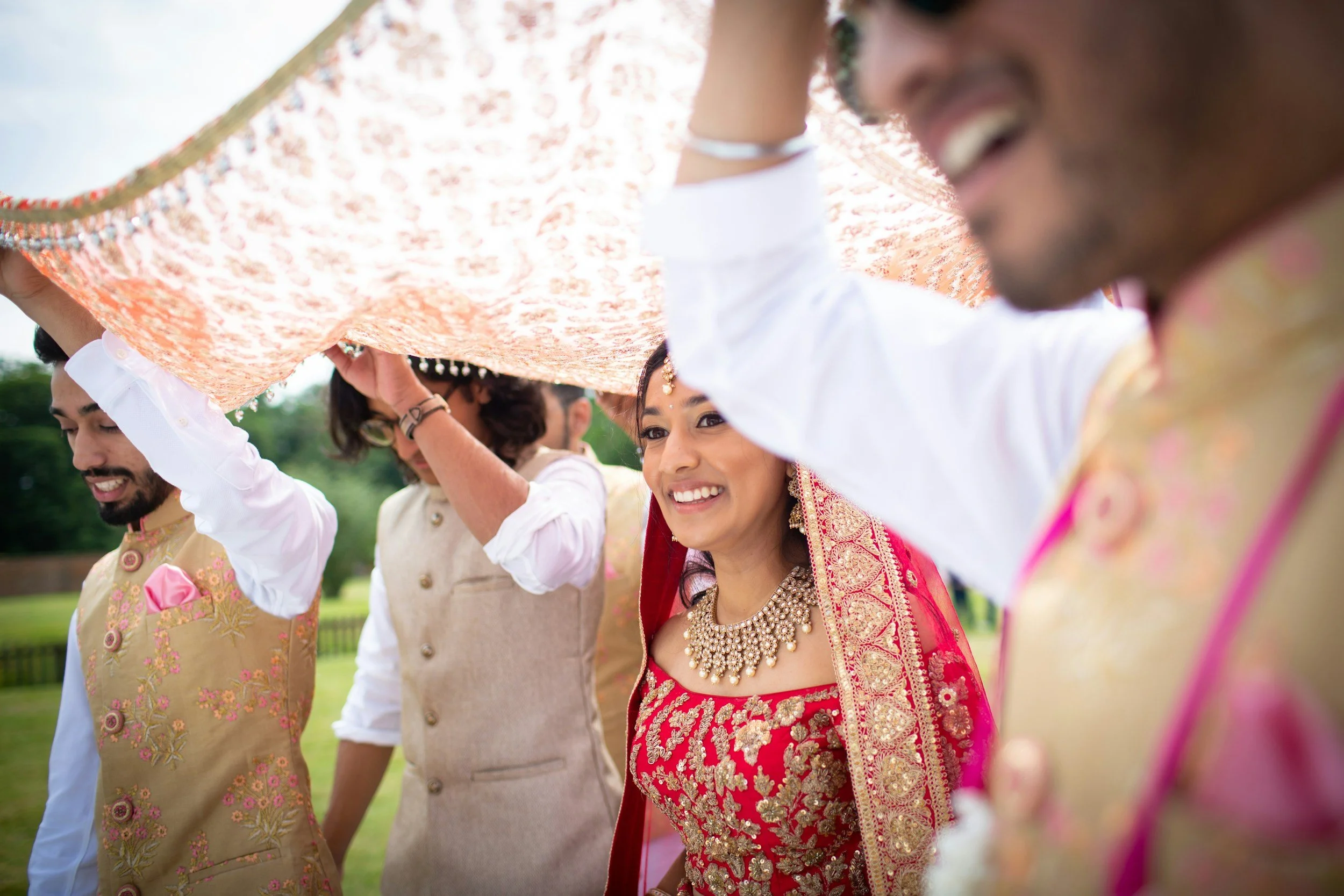 Group of people participating in an outdoor Indian wedding celebration, holding a decorated fabric canopy above their heads, with the bride wearing a red and gold traditional dress and jewelry smiling at the camera.