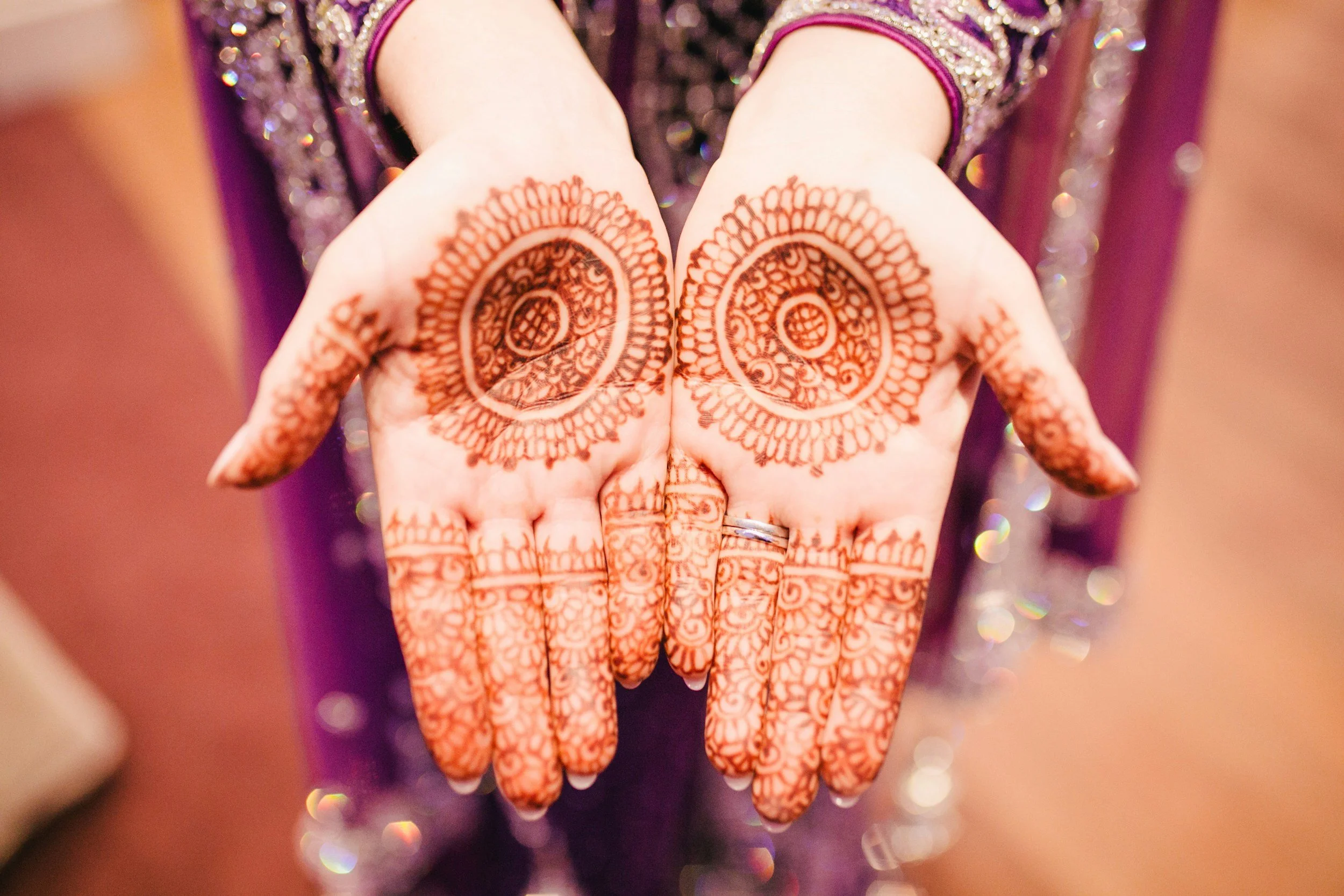 Close-up of hands with intricate henna designs on palms and fingers, wearing rings, with a blurred purple decorated background.