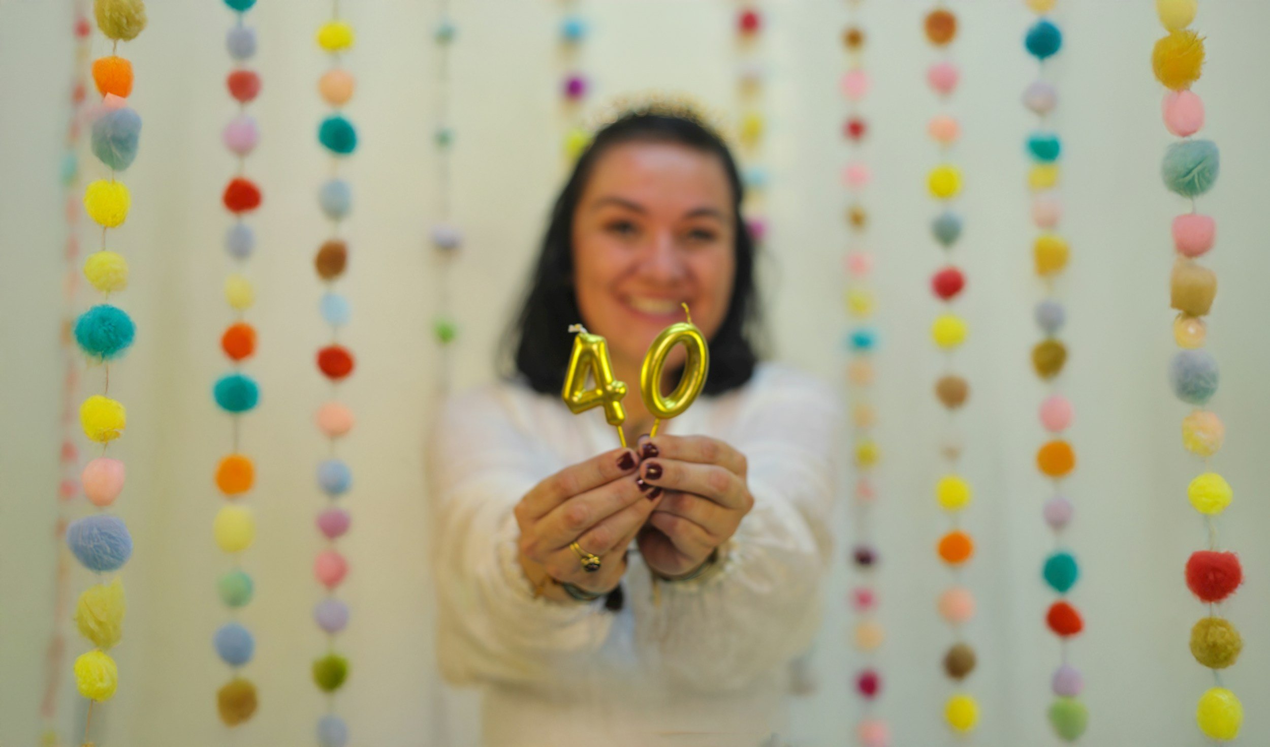 A woman holding gold balloons shaped as the number 40, smiling, with colorful felt pom-pom decorations hanging in the background.