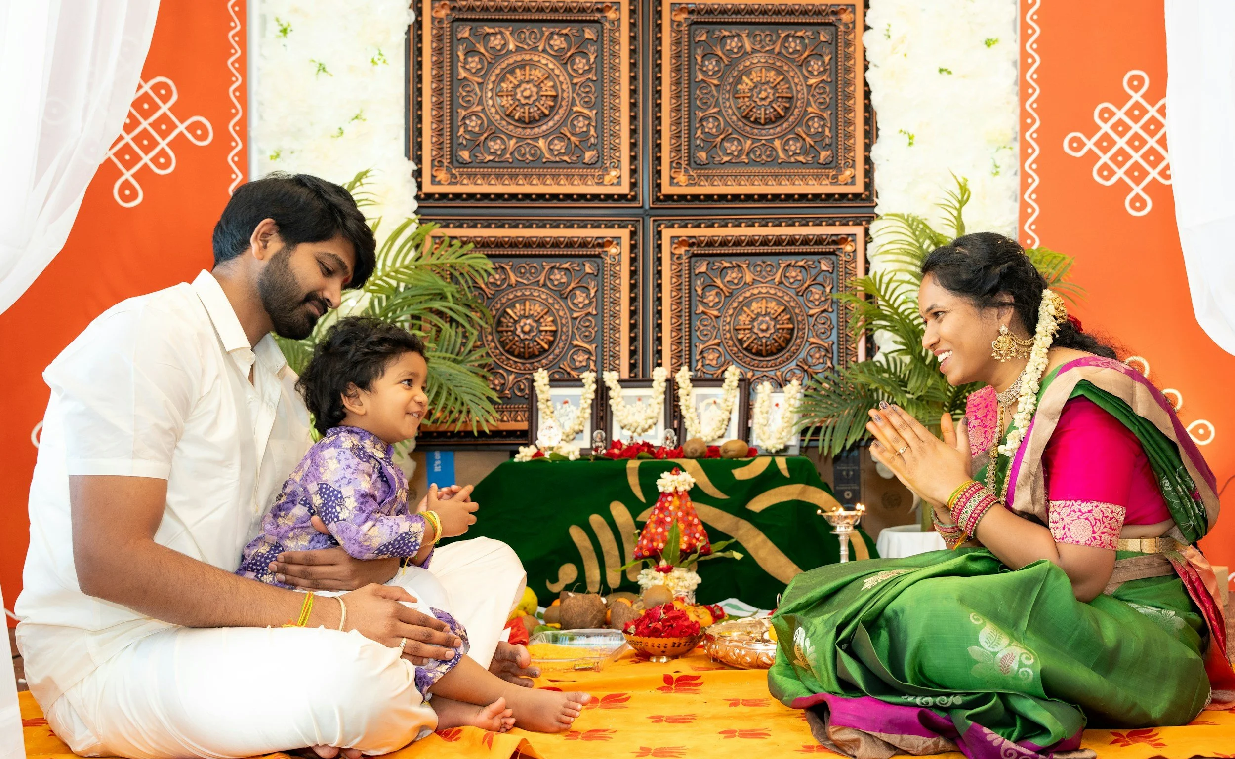 A family celebrating a traditional Indian religious or cultural ceremony indoors. A woman in a bright pink and green saree is smiling with folded hands, facing a man and a young boy. The man is sitting cross-legged, holding the boy on his lap. The boy is wearing a purple traditional outfit. There are ritual items, flowers, and incense on the floor and a decorated altar behind them with ornate wooden panels and white flower garlands.
