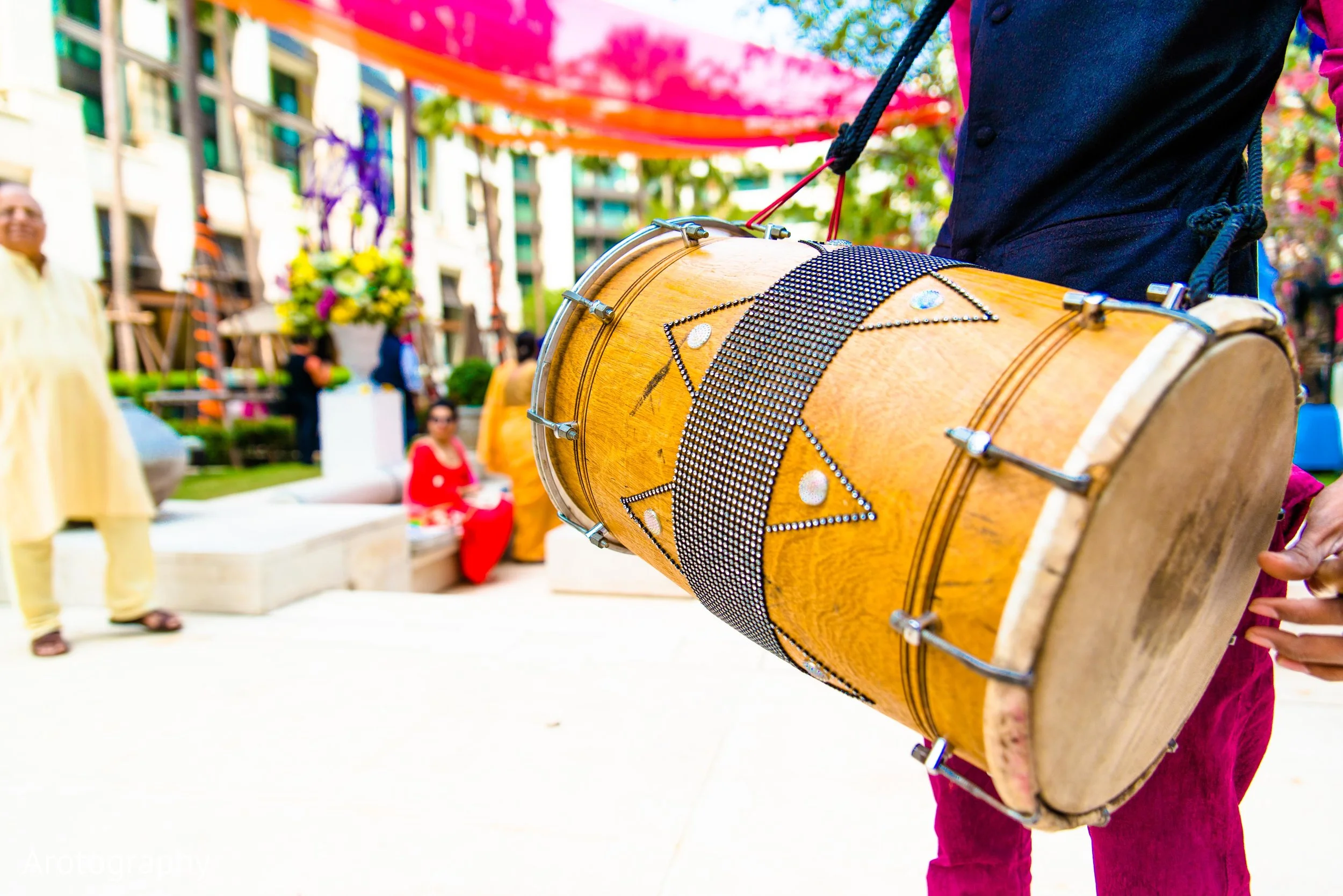 Close-up of a person holding a decorated wooden drum at an outdoor event with people seated and colorful decorations in the background.