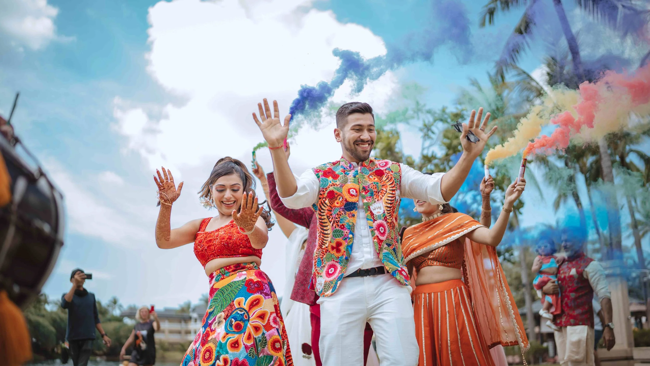 Group of people celebrating outdoors with colorful smoke sticks, dressed in traditional and vibrant clothing, smiling and dancing under a partly cloudy sky with trees in the background.