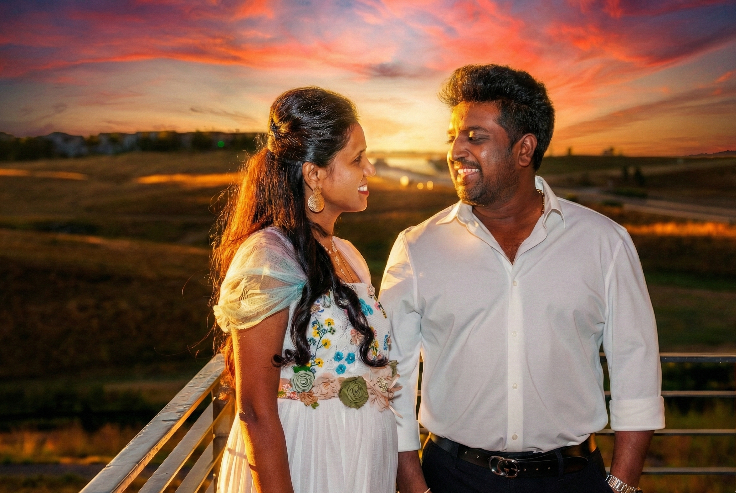A couple standing on a balcony at sunset, looking at each other and smiling. The woman is wearing a white dress with floral embroidery and earrings. The man is wearing a white shirt and black belt. The background features a colorful sky with shades of orange, pink, and purple, and a scenic landscape.