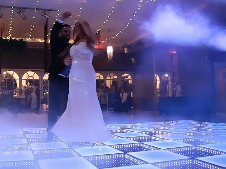 A couple dancing on an illuminated dance floor at a wedding reception with guests and decorative lighting in the background.