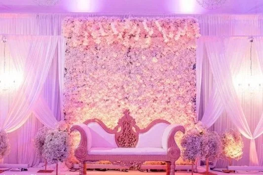 Elegant pink and white wedding stage with floral backdrop, ornate sofa, surrounded by flower arrangements and draped fabric
