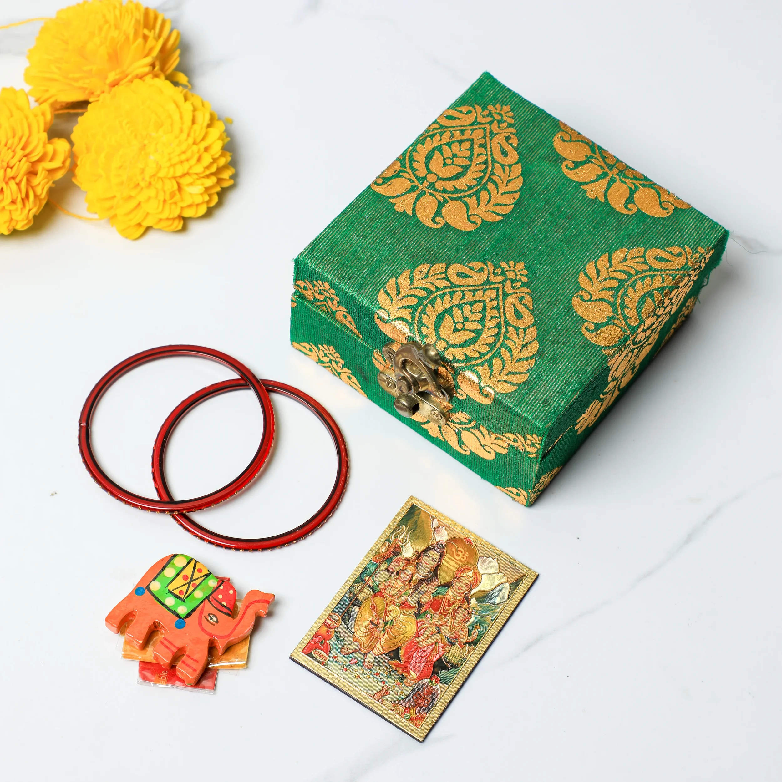 Decorative green box with gold patterns, yellow flowers, red bangles, a small colorful elephant figurine, and a picture of Hindu gods on a white surface.