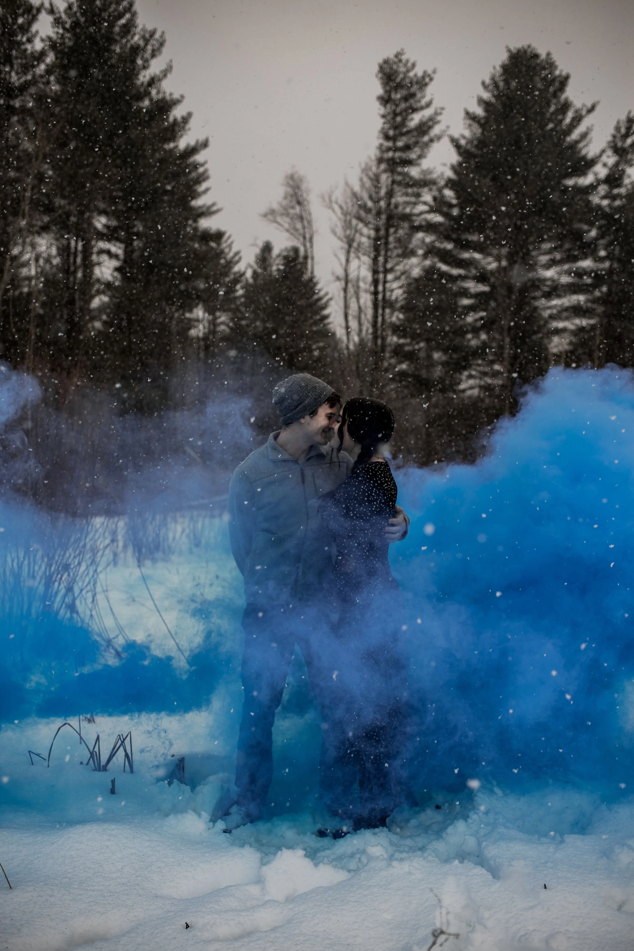 A couple stands close together in a snowy landscape, surrounded by blue smoke, with tall trees in the background.