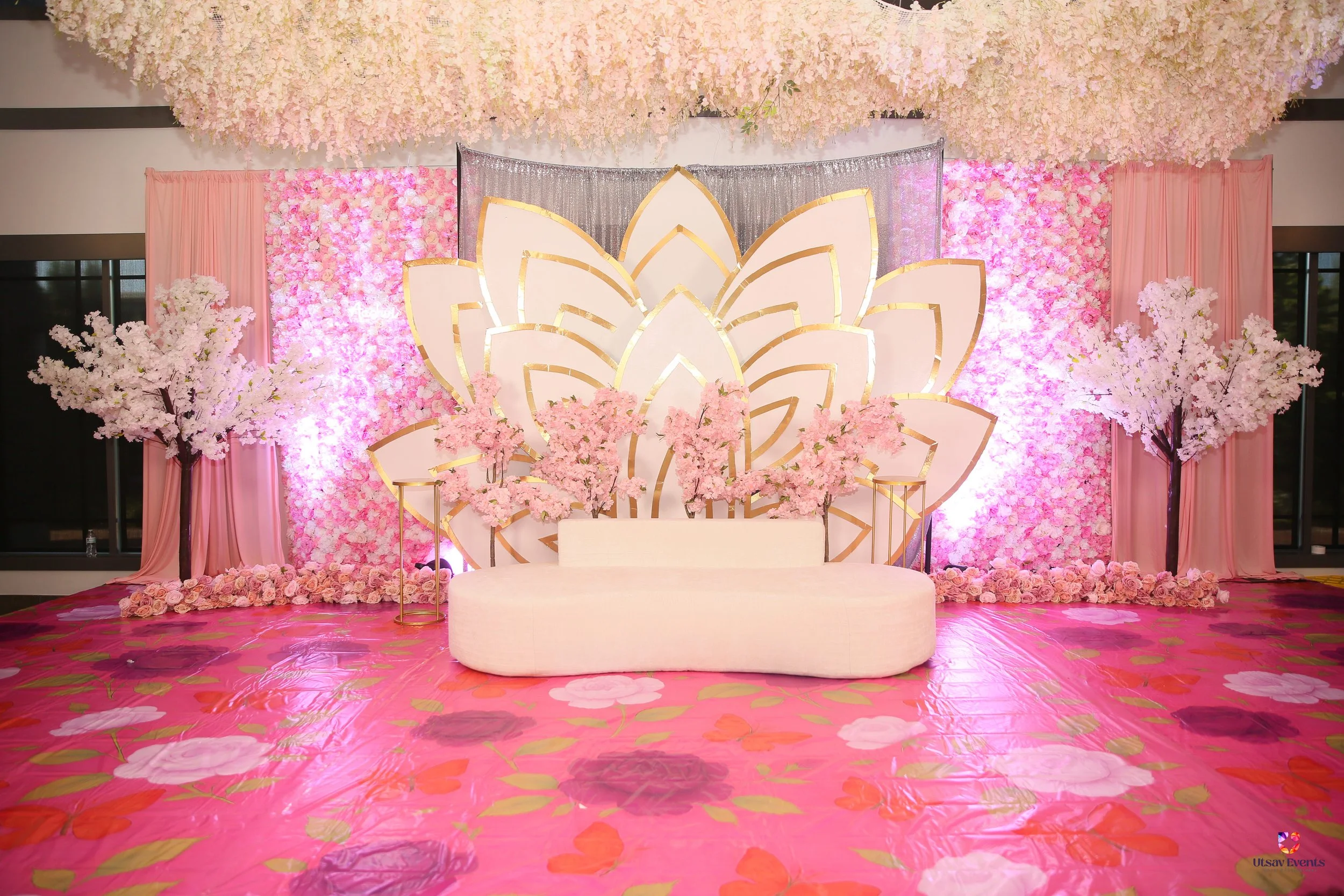 Elegant wedding stage decorated with pink and white flowers including cherry blossoms, a large stylized lotus flower backdrop with gold accents, pink drapes, and floral carpeted flooring.