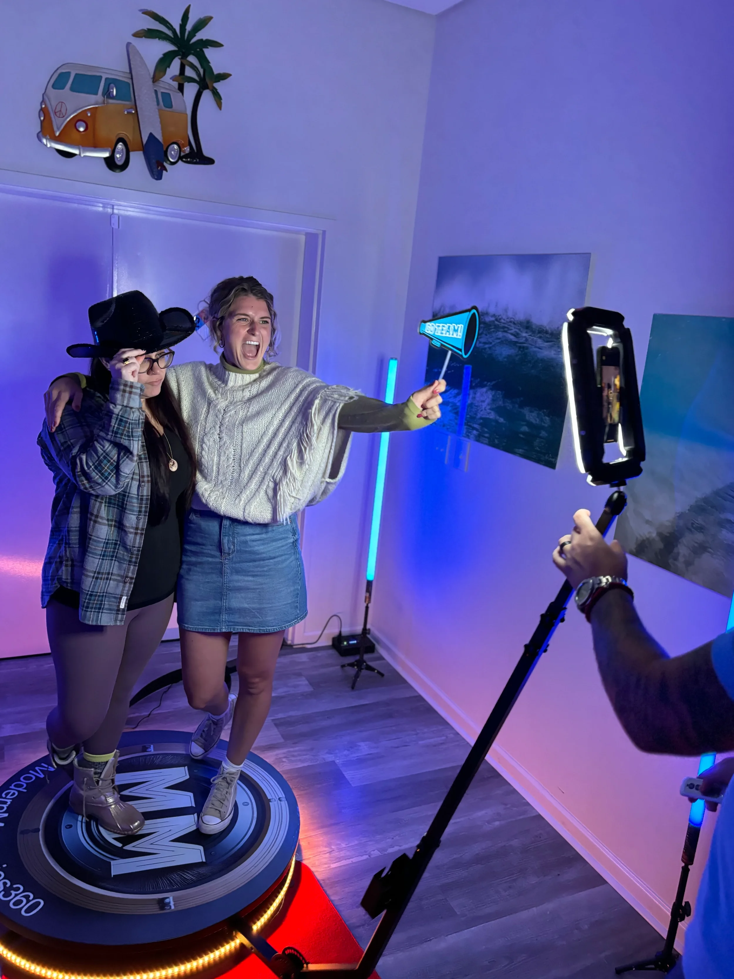 Two women posing for a photo on a circular platform with a camera and a person taking their picture, in a room with neon lights and ocean pictures on the wall.