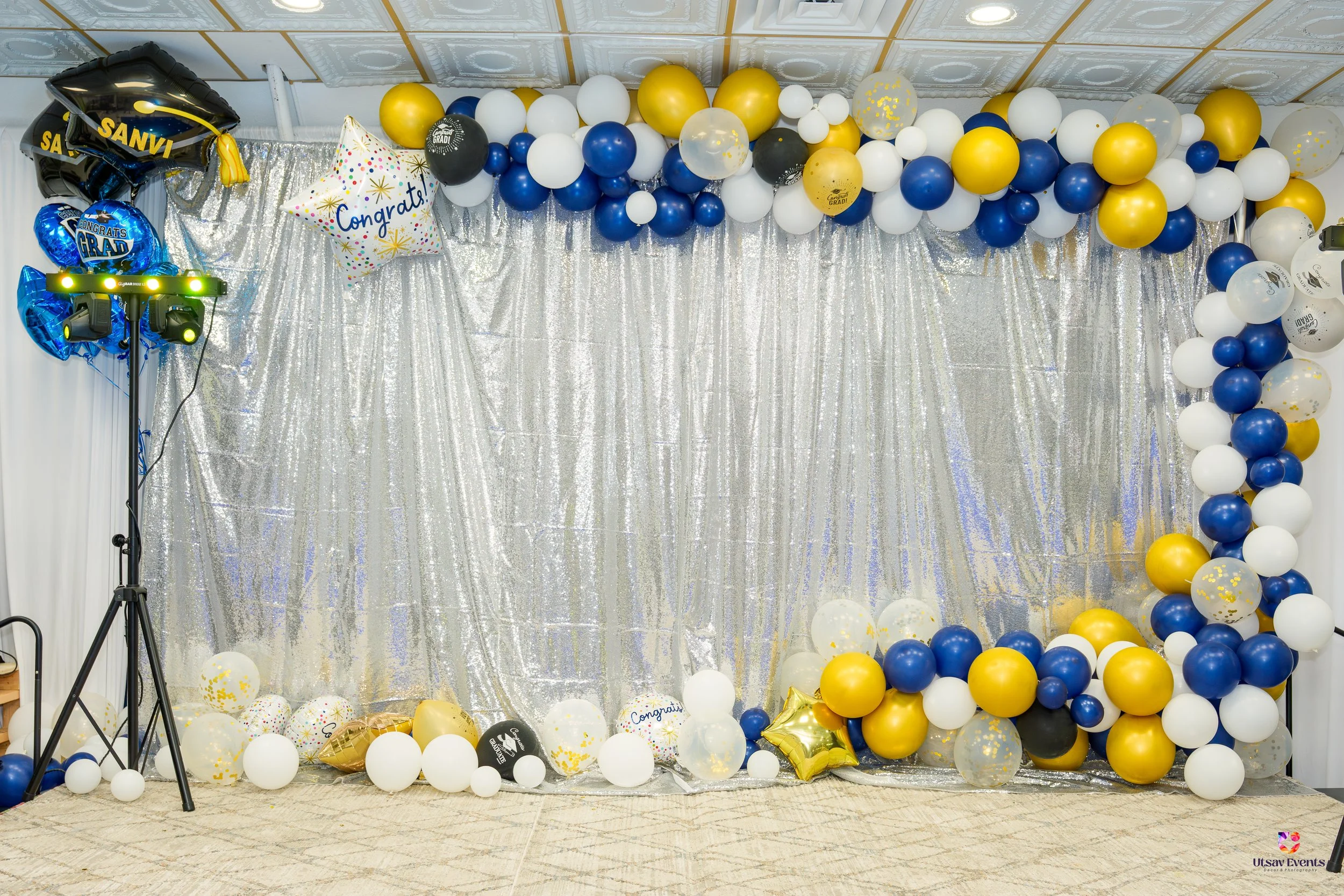 Decorative backdrop with a balloon arch and balloons on the floor for a graduation celebration. The balloons are in white, navy blue, gold, and black. There are metallic curtains background and a star-shaped balloon with 'Congrats' written on it. A balloon with 'Grad' written on it and a graduation cap balloon are also part of the decor.