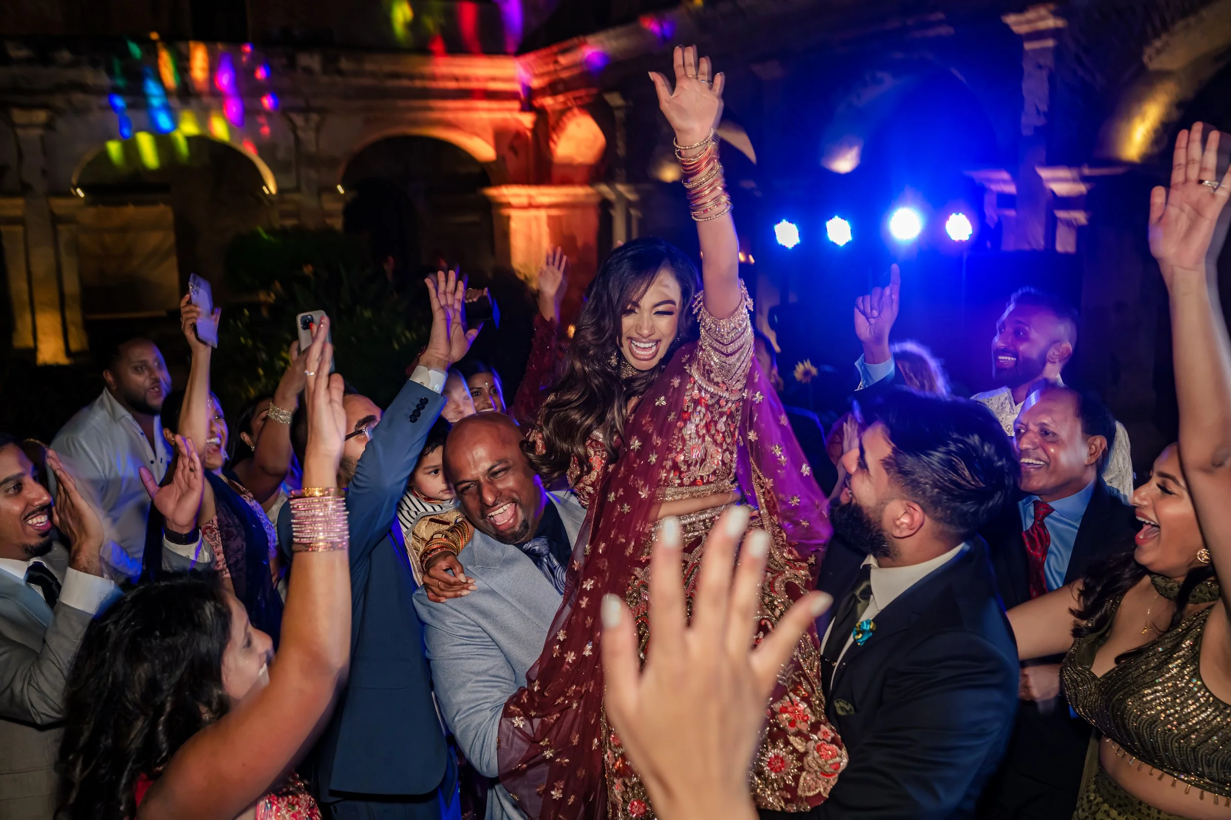 A group of people celebrating at a party, with a woman in traditional Indian dress being lifted up, surrounded by smiling, dancing guests in formal attire, colorful lights, and a historic building in the background.