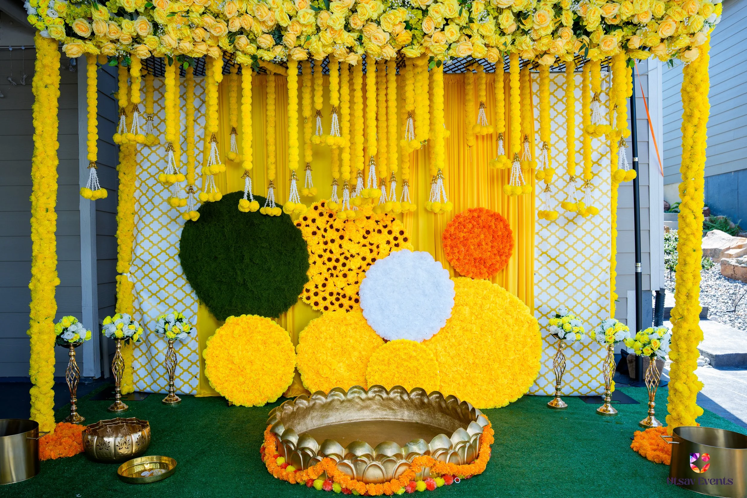 Decorative floral setup featuring a yellow theme with hanging yellow pom-poms, yellow roses, and large shaped floral arrangements in yellow, orange, green, and white. Includes a gold tray with marigold flowers at the base, set on a green grassy surface.