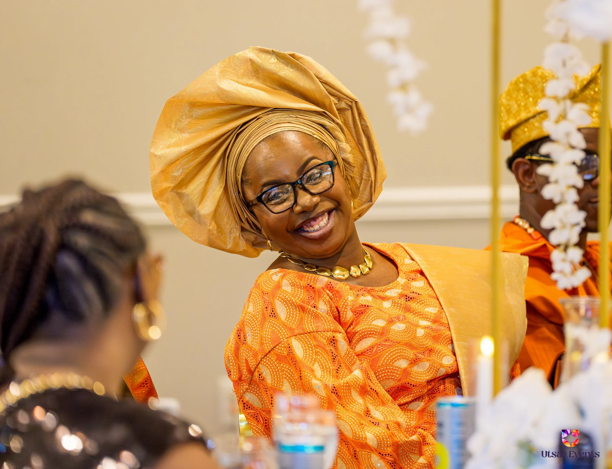 A smiling woman wearing glasses, traditional African attire in gold and orange, and a matching head wrap, at a celebration or event with decorated table and other guests in the background.
