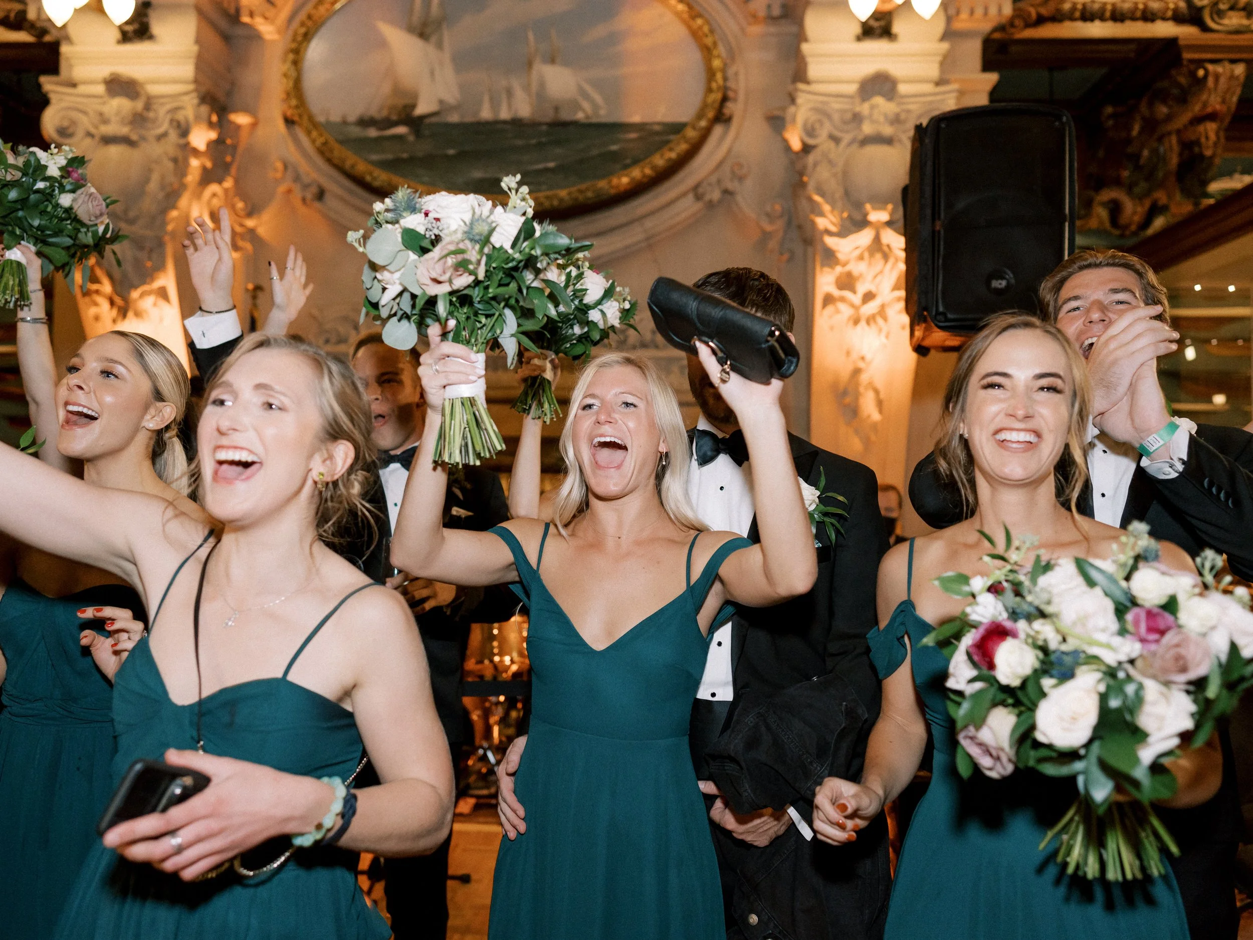 A group of joyful wedding guests dressed in formal attire, celebrating and raising their arms, some holding bouquets, inside a decorated ballroom with ornate artwork and a large painting of ships in the background.