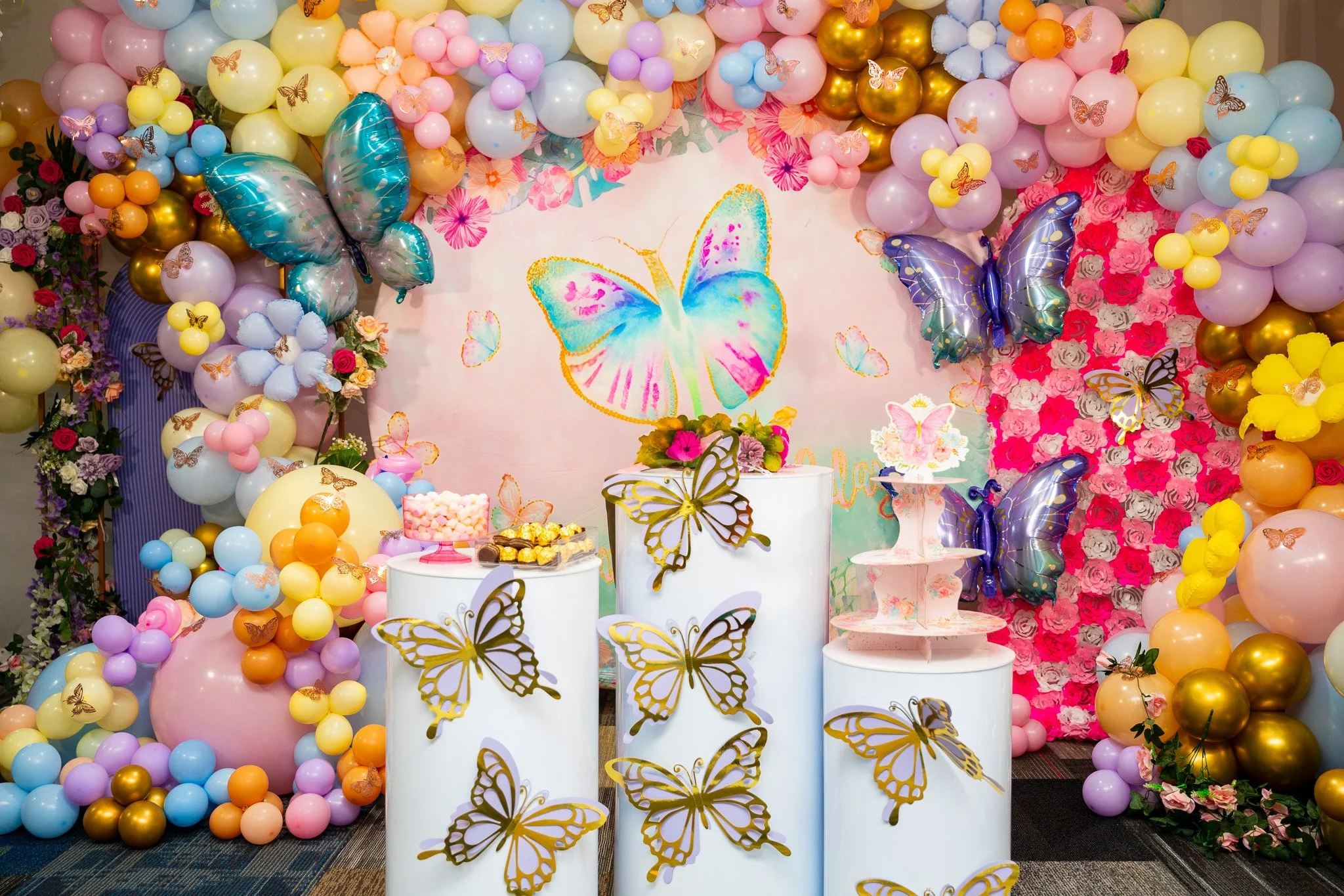 Decorative butterfly-themed birthday or celebration backdrop with pastel balloons, butterfly cutouts, and floral arrangements.