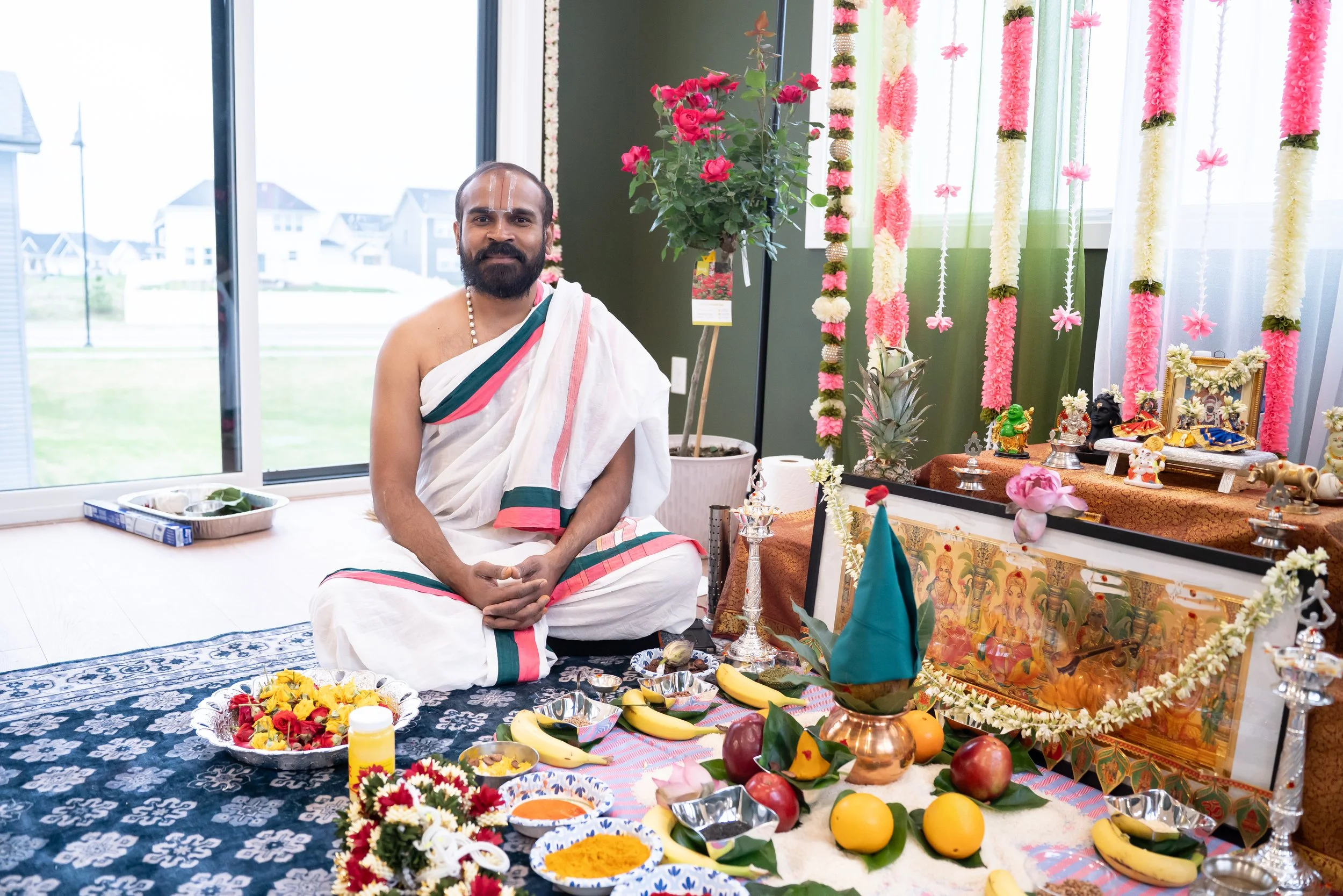 A man dressed in traditional Hindu religious attire sitting on a decorated floor during a religious ceremony, with prayer items, fruits, flowers, and deity images around him, in a room decorated with flowers and garlands.